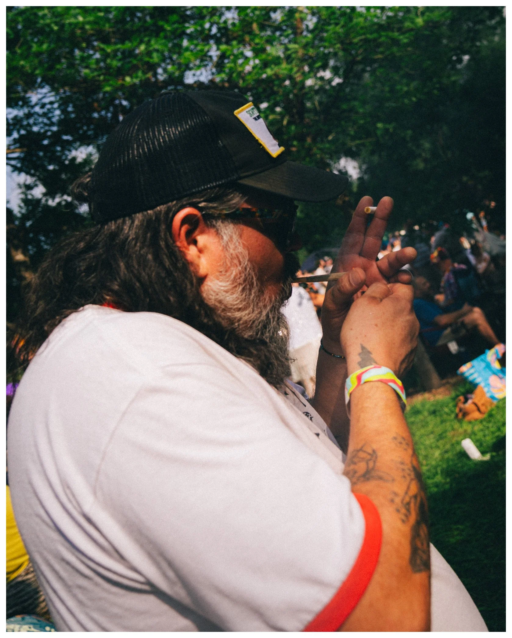 A man with long hair and a beard wearing a black mesh cap, sunglasses, and a white t-shirt with orange trim, holding a lit cigarette, with tattoos on his arm and wristbands, outdoors near green trees and other people in the background.