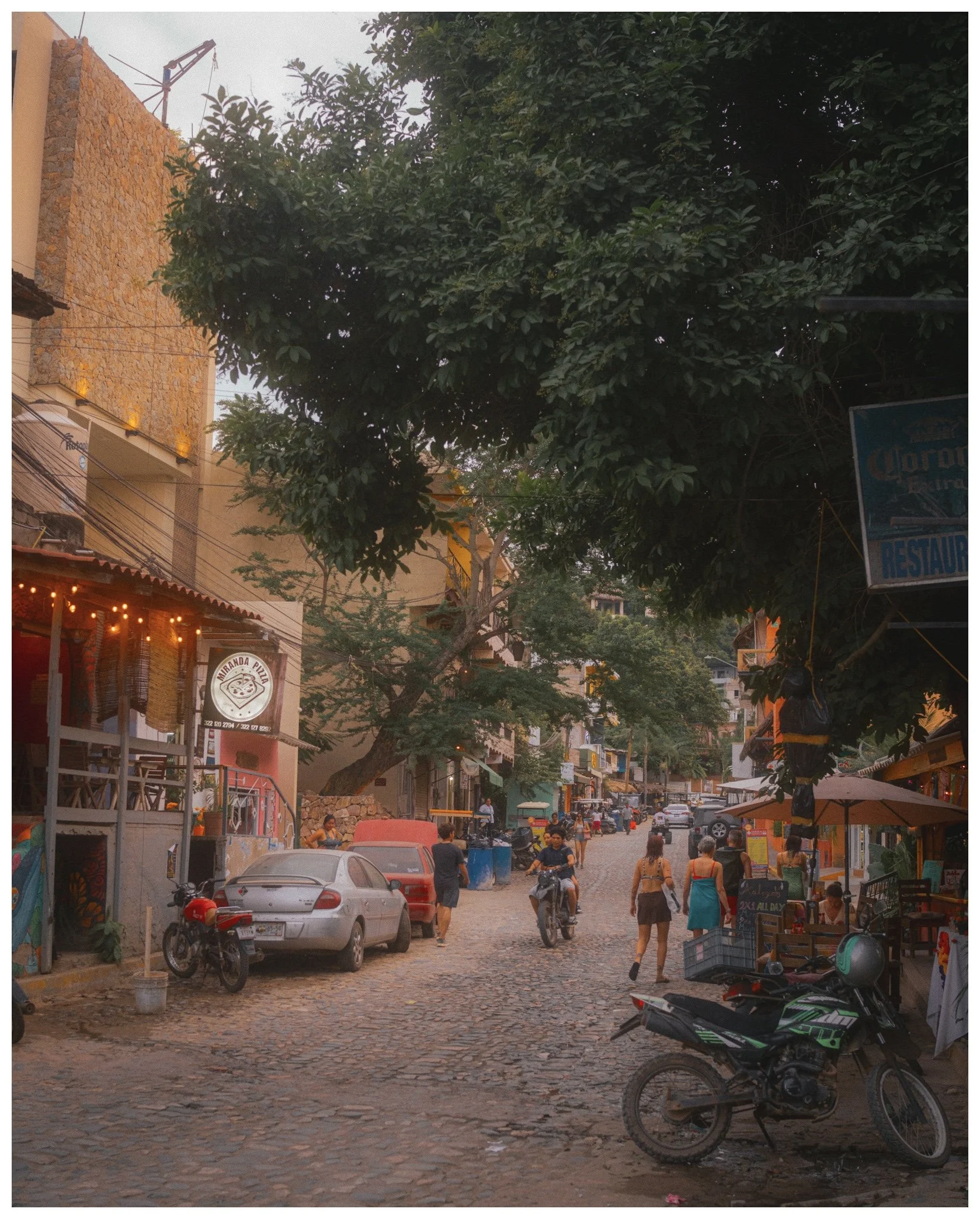 A busy cobblestone street scene in an urban area with parked cars and motorcycles, pedestrians walking, storefronts and signs, large trees lining the street, and buildings with warm-colored facades.