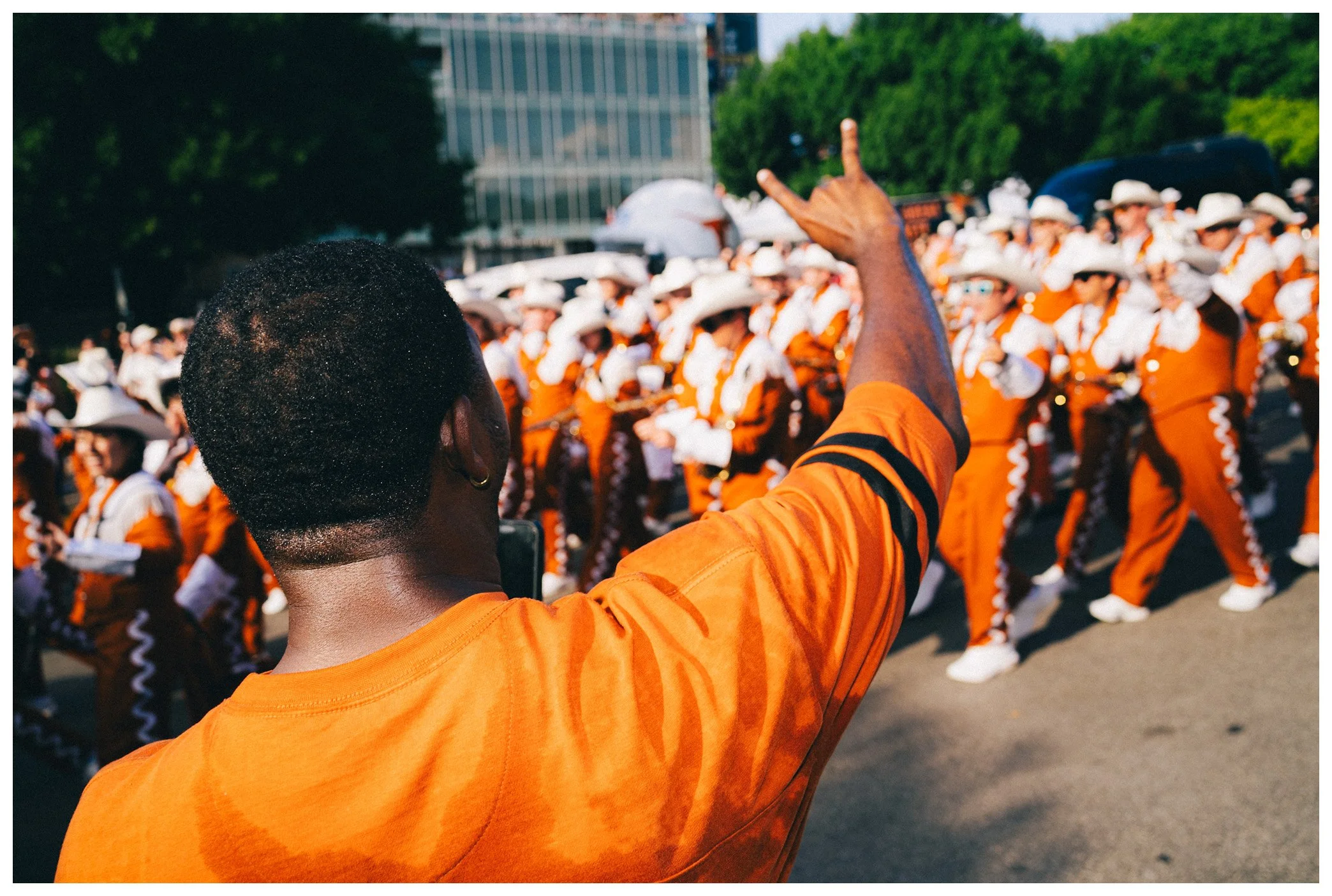 A parade with people dressed in orange and white marching band uniforms, many with white cowboy hats, as a man in an orange shirt waves in the foreground.