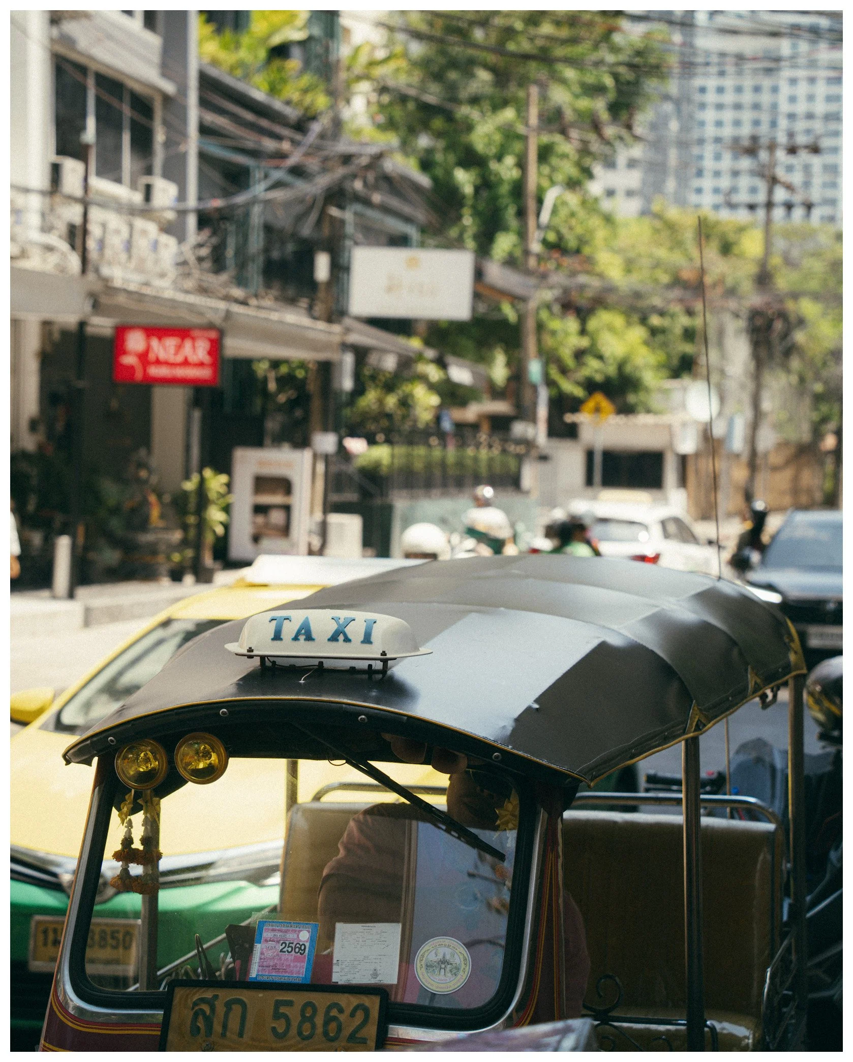 A tuk-tuk or auto rickshaw with a taxi sign on the roof, parked on a busy city street with other vehicles and buildings in the background.
