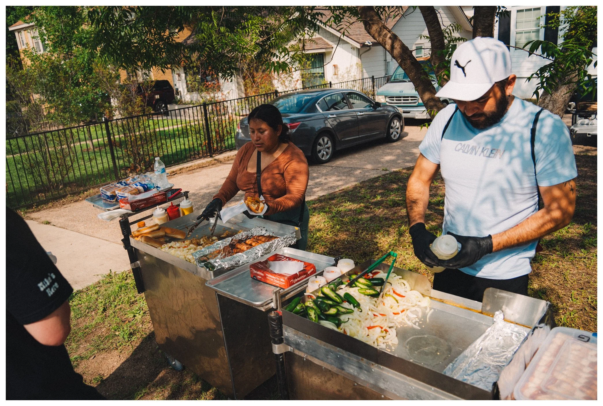 People preparing food at an outdoor barbecue with parked cars and houses in the background.