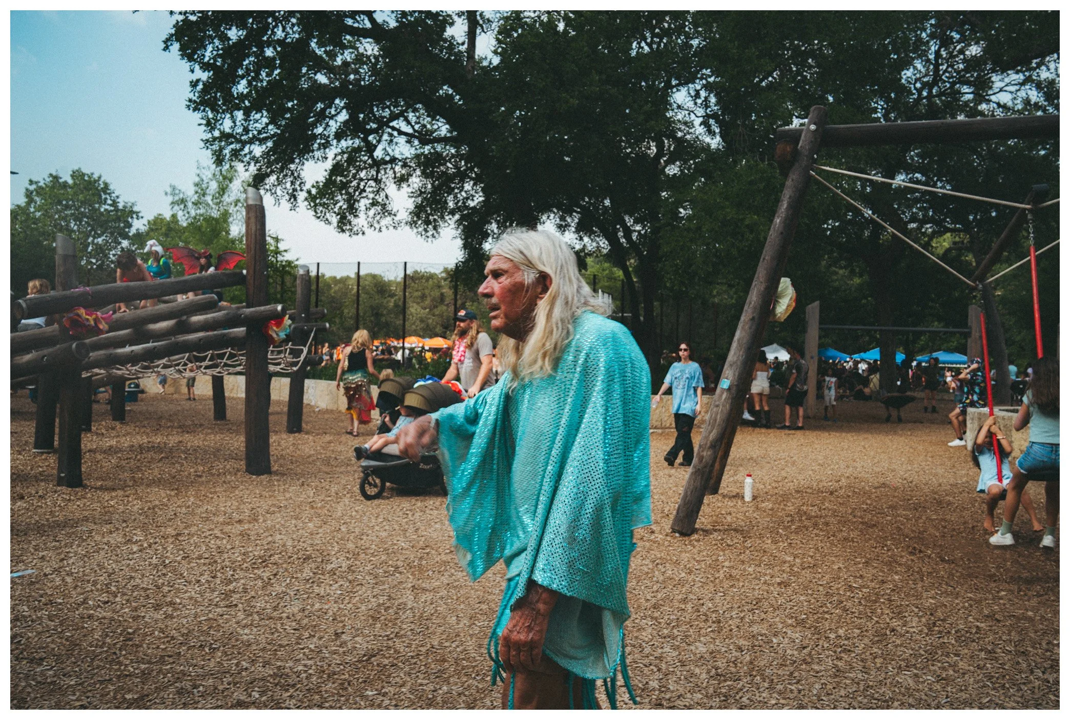 An elderly woman with long white hair wearing a turquoise shawl stands at a playground during daytime, with children and adults in the background, some under umbrellas, and trees surrounding the area.