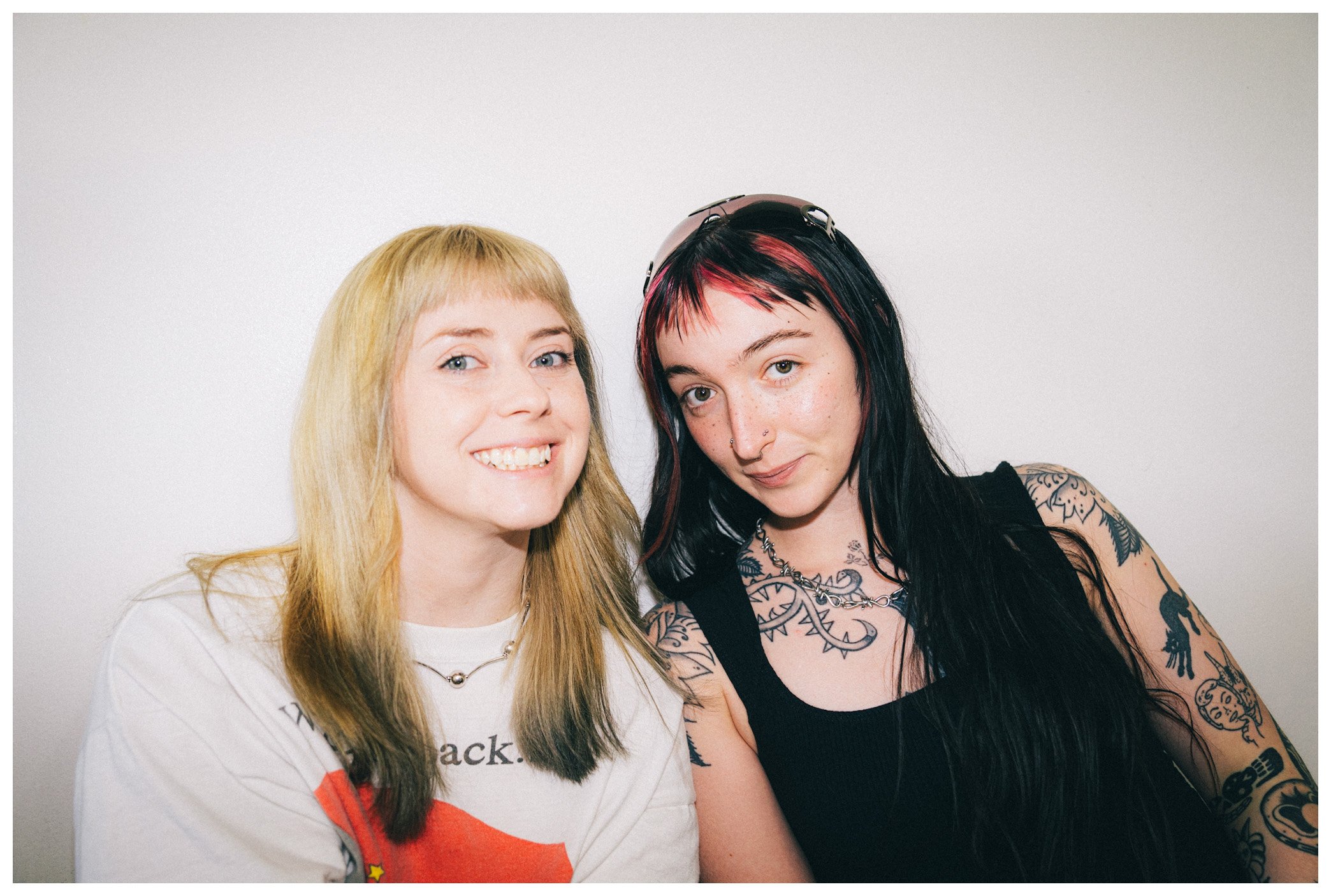 Two women posing for a photo against a plain white wall. The woman on the left has long blonde hair, blue eyes, and is smiling. The woman on the right has black hair with red highlights, multiple piercings, tattoos on her arms and chest, and is makin