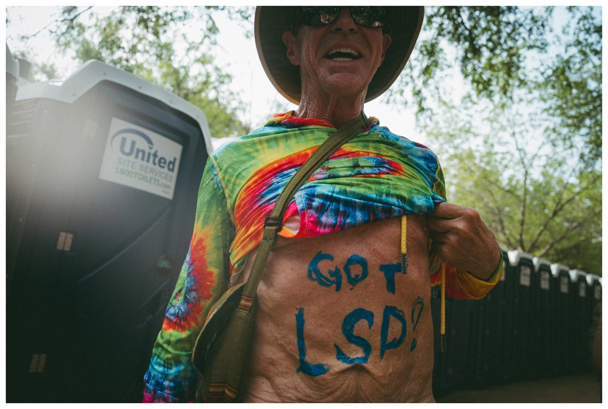 Close-up of a man wearing a tie-dye shirt, a wide-brimmed hat, and sunglasses outdoors, revealing painted messages on his chest, with trees and a fence in the background.
