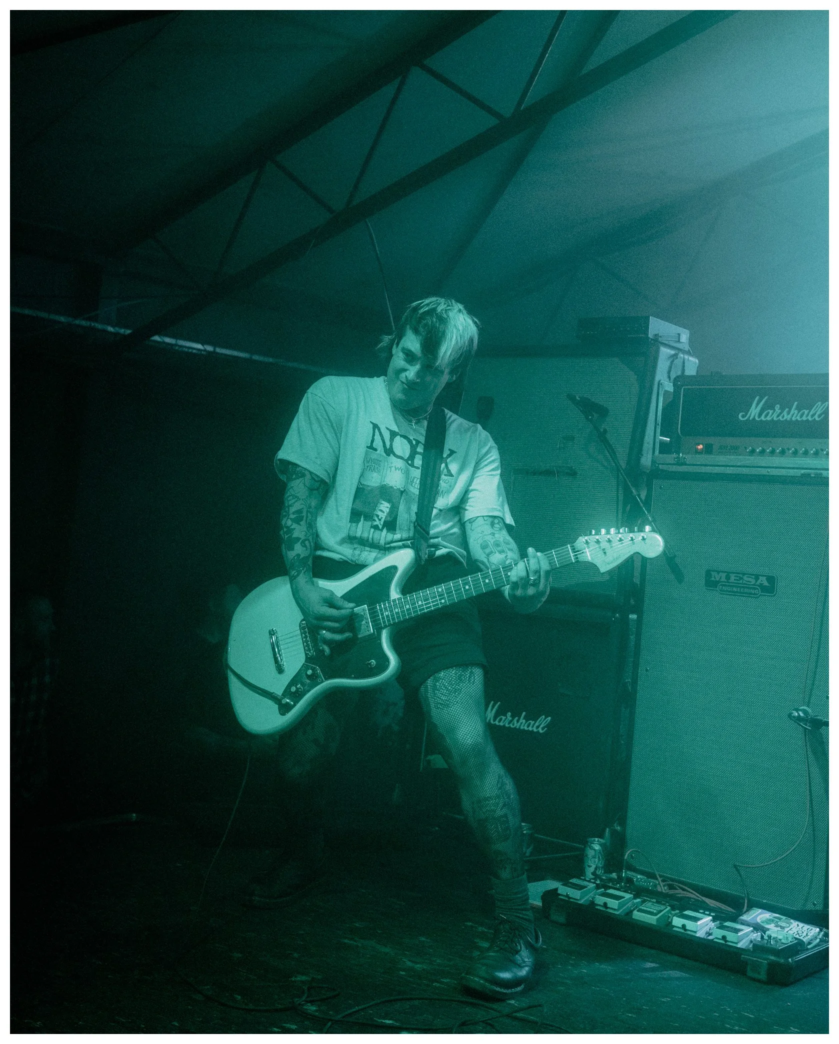 A musician playing an electric guitar on stage with Marshall amplifiers behind him and guitar effect pedals on the floor. The stage is dimly lit with teal-colored lighting.