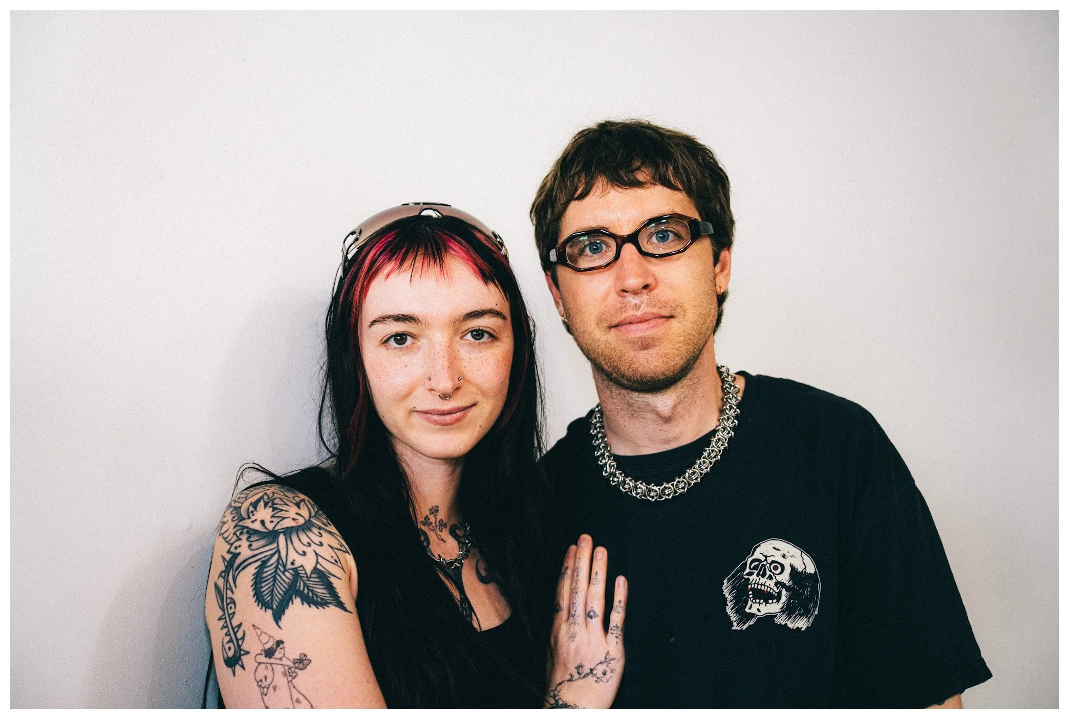 A young woman with red and black hair, tattoos on her arm and chest, and piercings on her face, standing next to a young man with glasses, short brown hair, and a chain necklace. They are both wearing black shirts and posing against a plain white wal