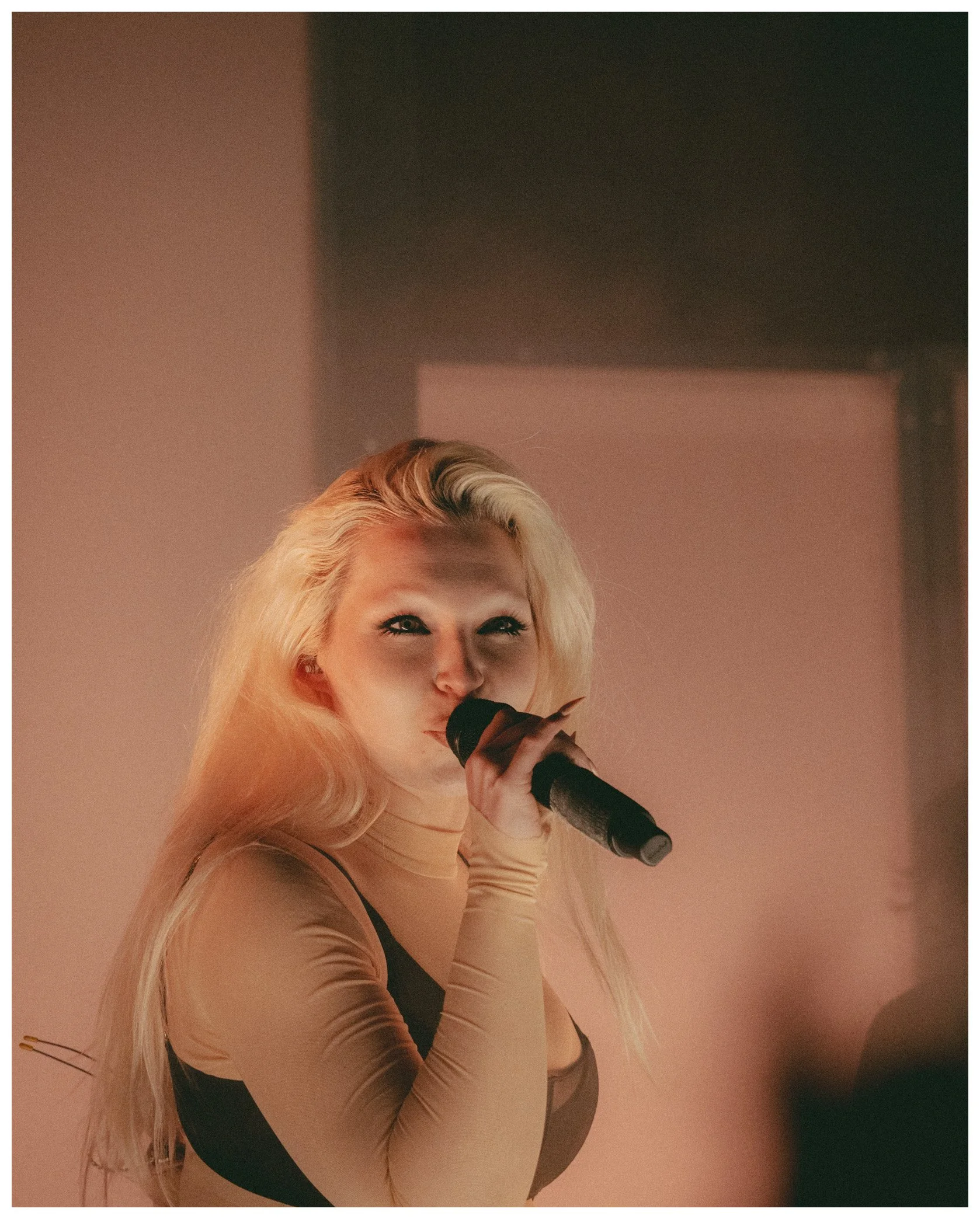 A woman with long blonde hair and bright makeup is holding a microphone, looking towards the camera, with a pink backdrop.