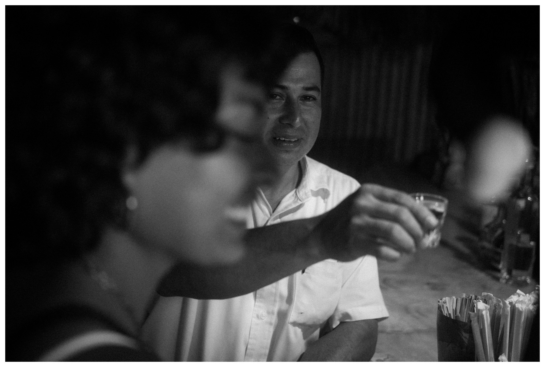 A man in a white shirt smiling and holding a drink at a social gathering, with a group of people and food on the table in the background.