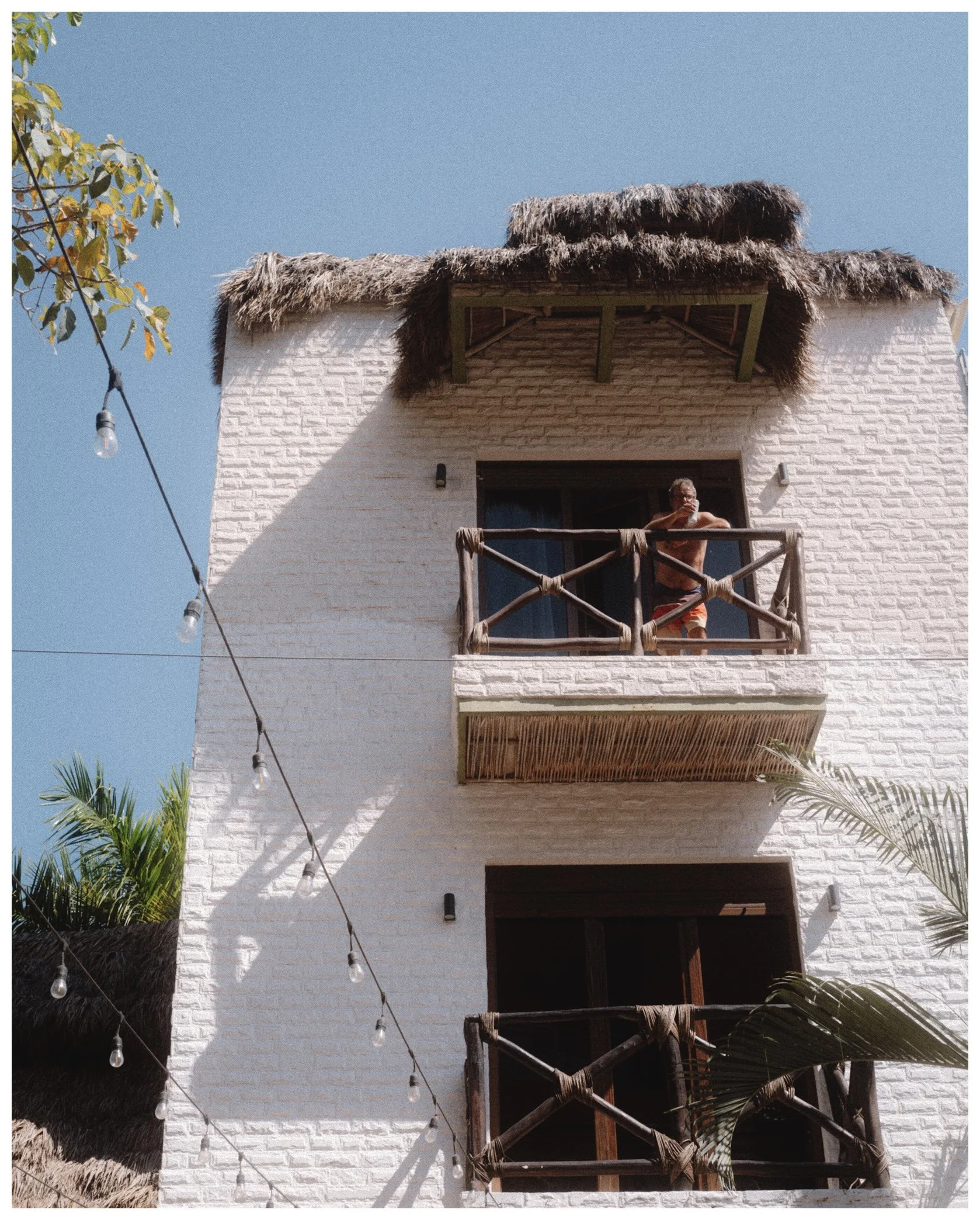 A multi-story white building with thatched roofing over the balconies, located in a tropical setting. A person is standing on the second-floor balcony, drinking from a cup. There are string lights hanging across the image and tropical palm trees visi