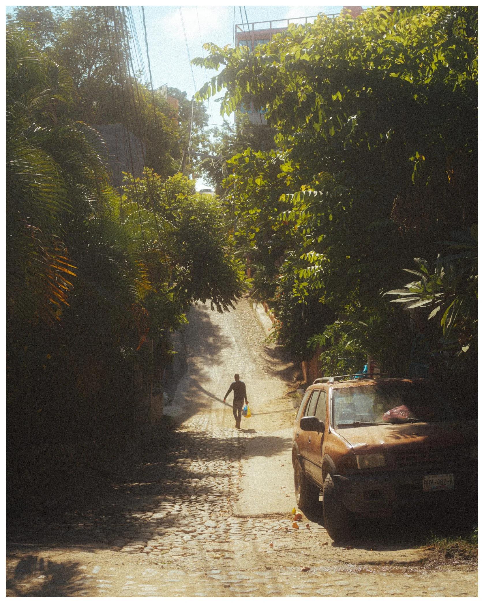 A cobblestone street with a parked brown car on the right, a person walking away carrying a yellow bag, shadows from trees on the street, and lush green foliage and trees lining the street on both sides, with a building and power lines in the backgro
