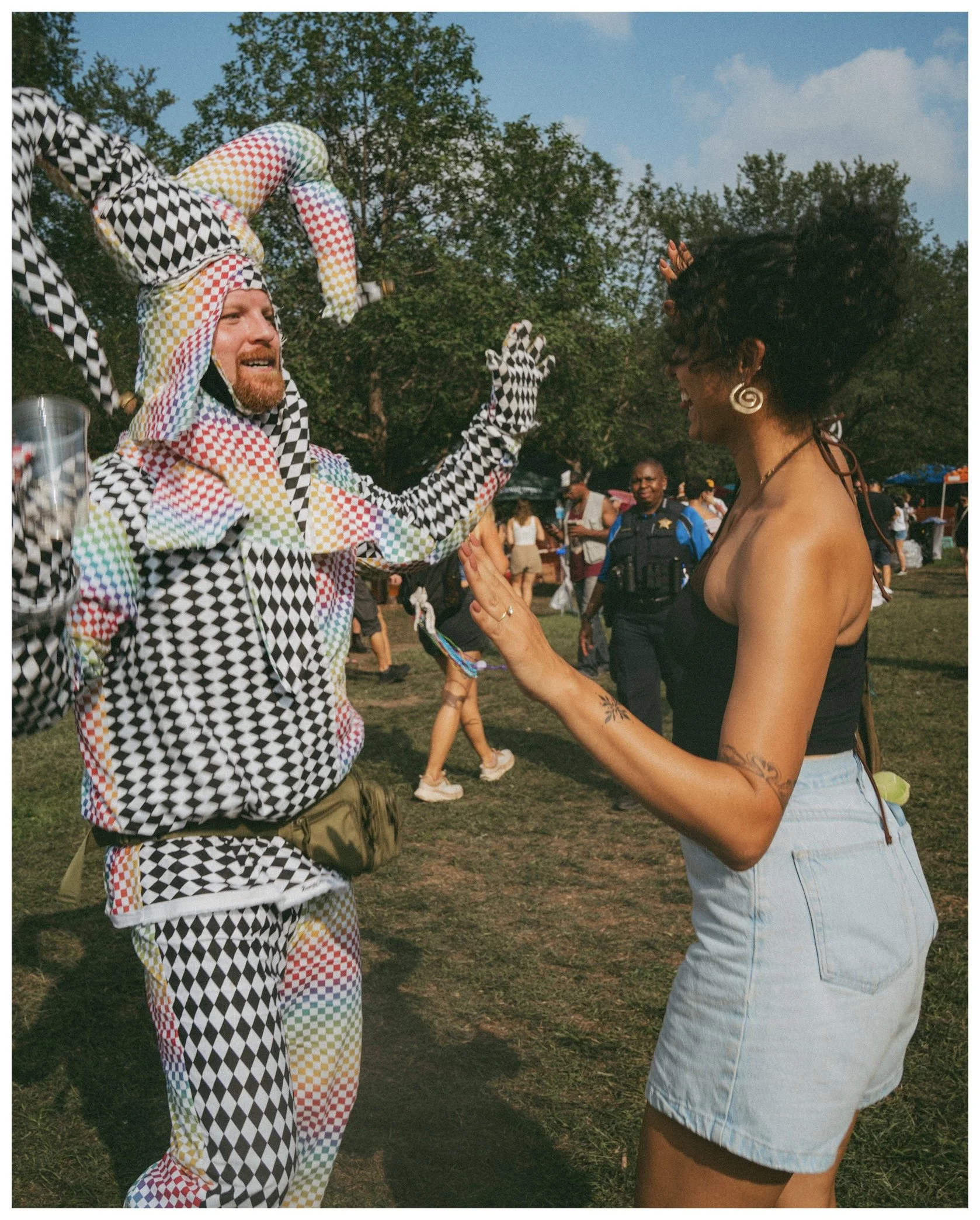 A man wearing a multicolored checkered jester costume and a woman with curly hair, a tattoo, and earrings engaging in a handshake or greeting at an outdoor event, with other people and trees in the background.