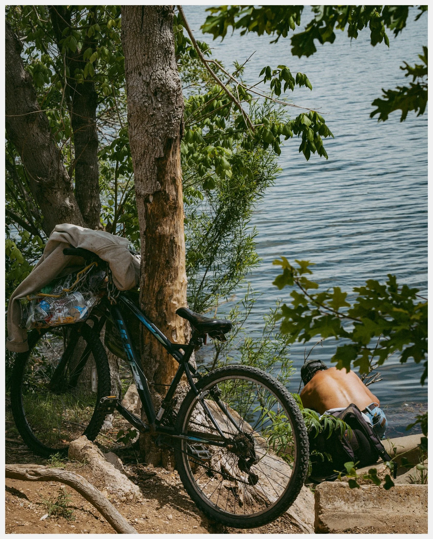 A bicycle parked near a tree by a body of water, with a backpack and water bottles attached to the bike, and a person lying on the rocks near the water wearing a hat.