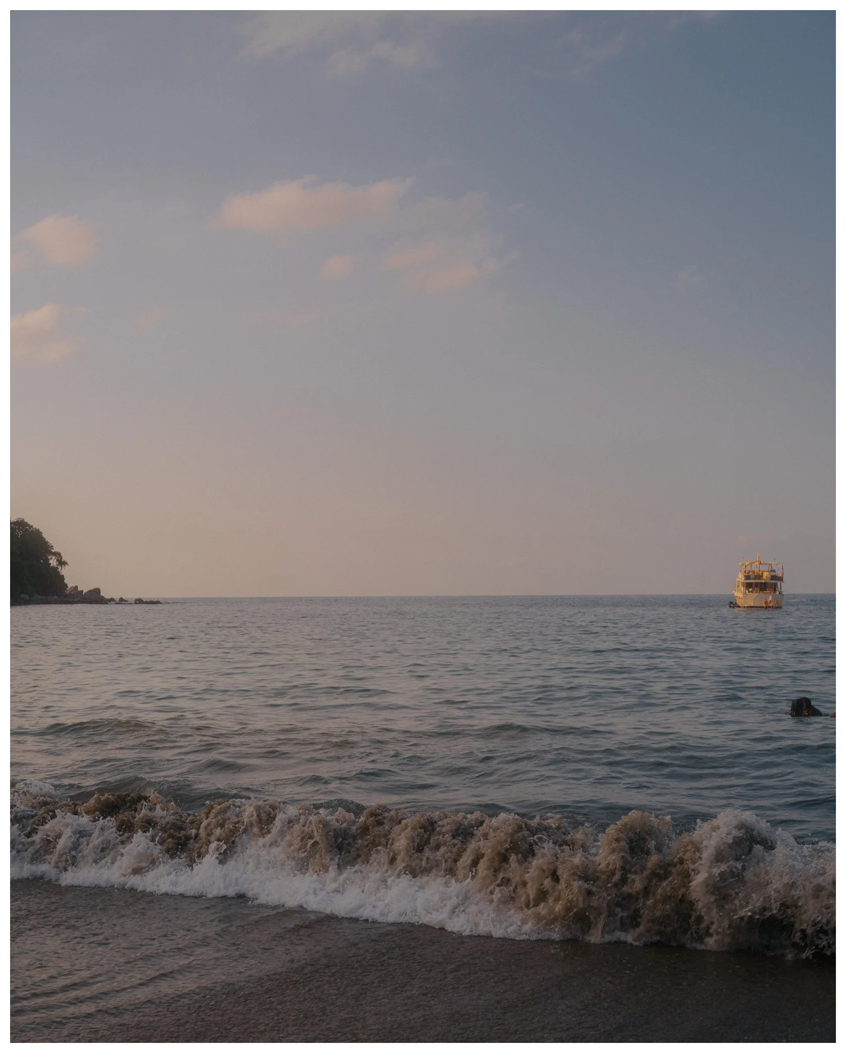 View of a beach at sunset with waves crashing on the shore, a palm tree and some rocks on the left, and a boat on the water on the right.