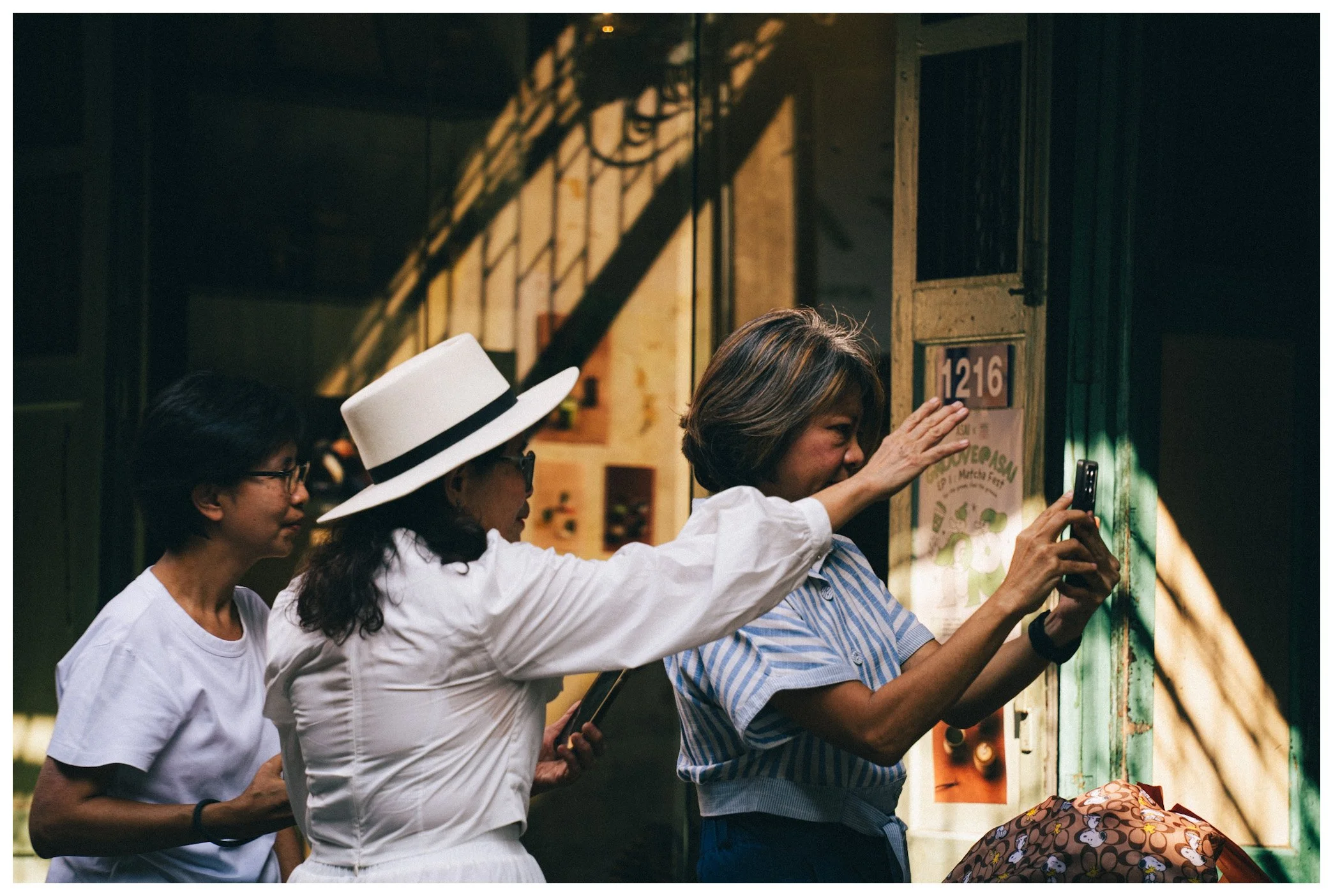 Three women with dark hair, wearing white and striped shirts, standing in line at an indoor food or drink stall, with one woman taking a photo of a menu or display with her phone.