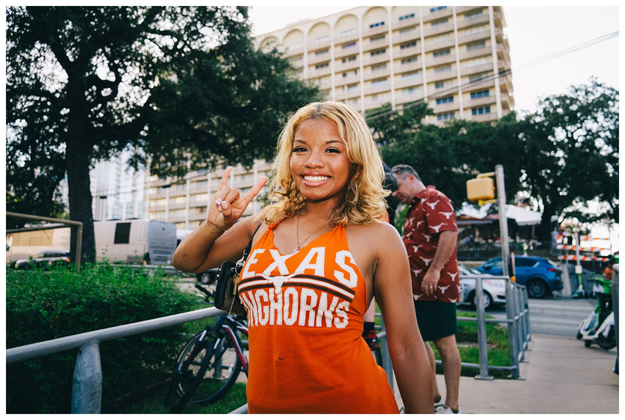 A young woman with blonde hair, smiling and making a peace sign with her right hand, wearing an orange Western Athletic Conference Texas Tech shirt. She is standing outdoors near a park with trees, a high-rise building, and people in the background.