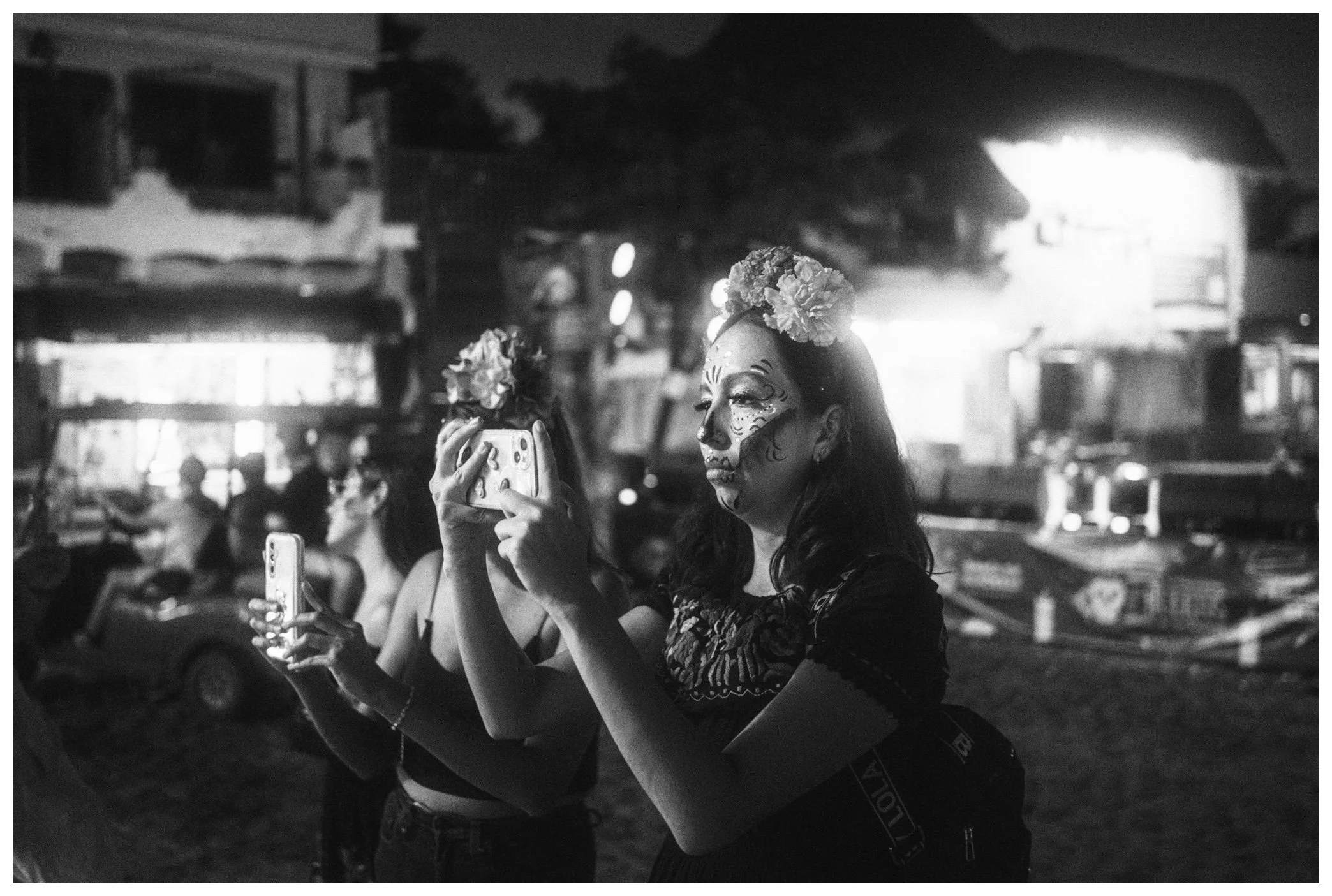 A woman with face paint and flowers on her head taking a selfie with a phone at a night event, with other people in the background.