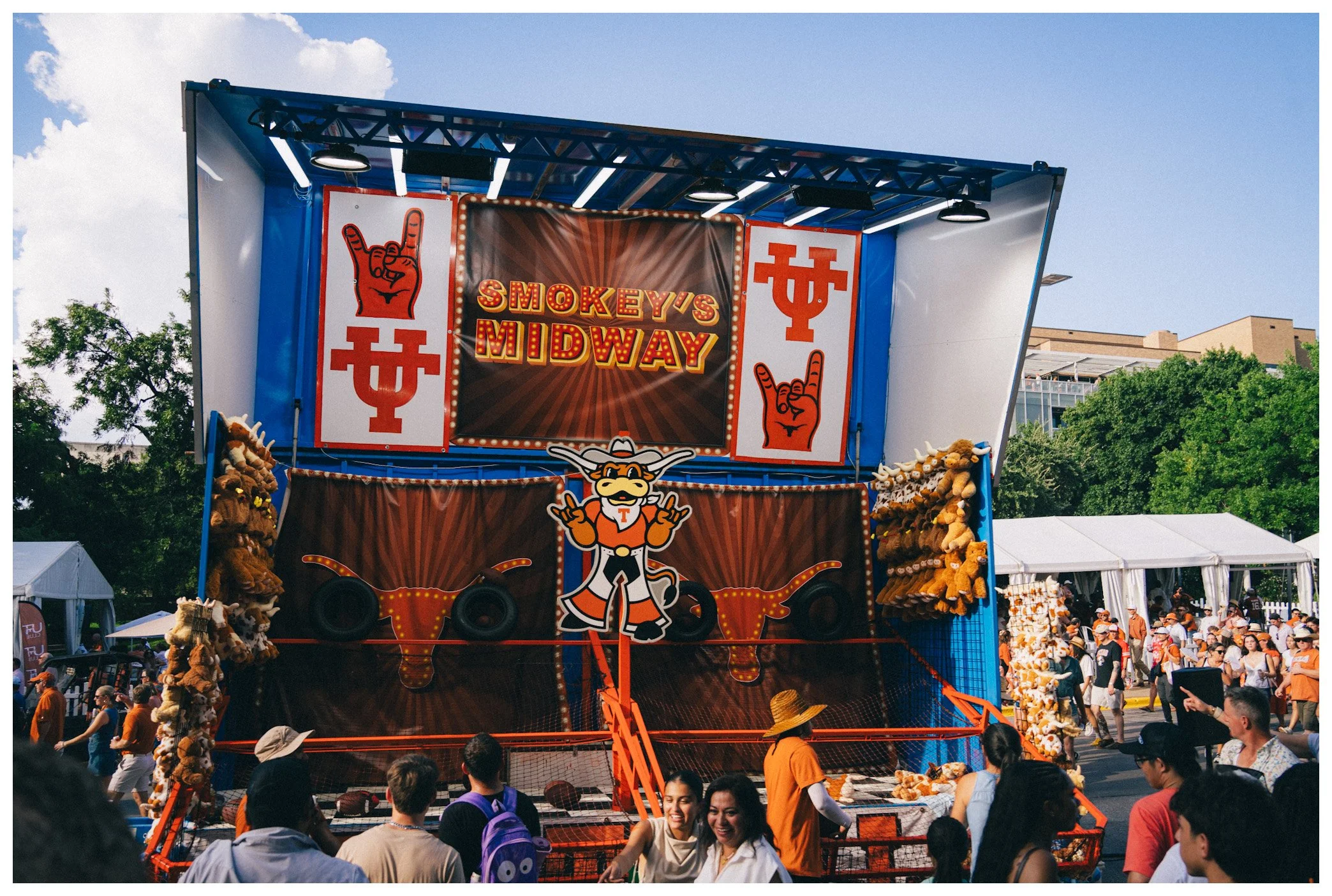 People gathered around a carnival midway with a booth named Smokey's Midway, decorated with cartoonish cowboy and casino-themed graphics, and plush teddy bears as prizes.