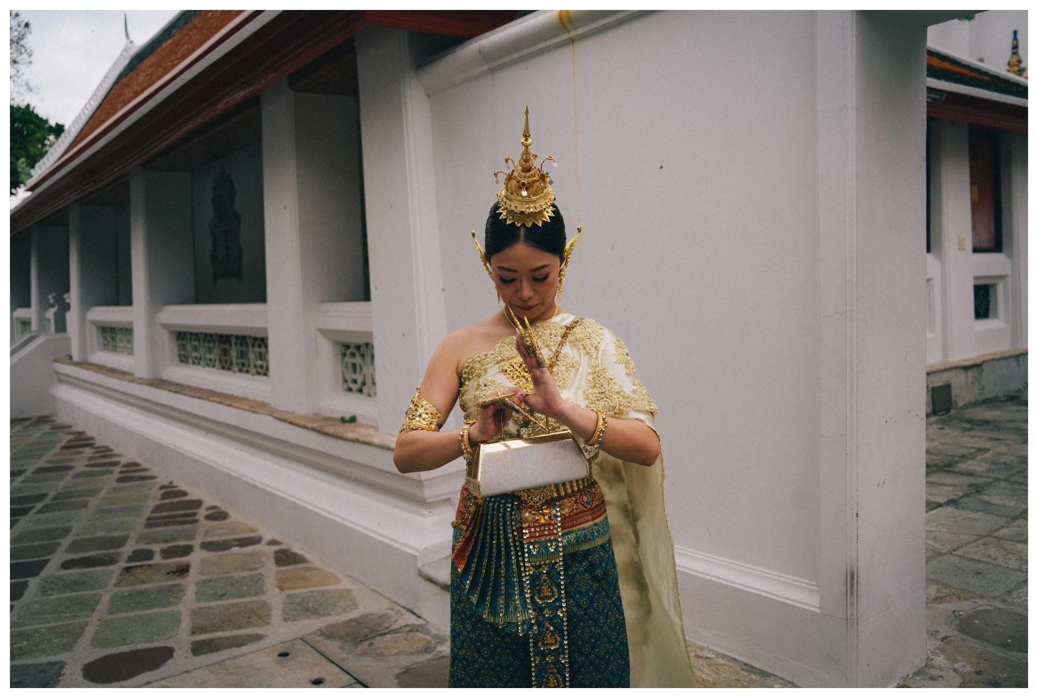 A woman dressed in traditional Thai attire, holding her hands in a prayer position, standing outside a white building with a tiled pathway.