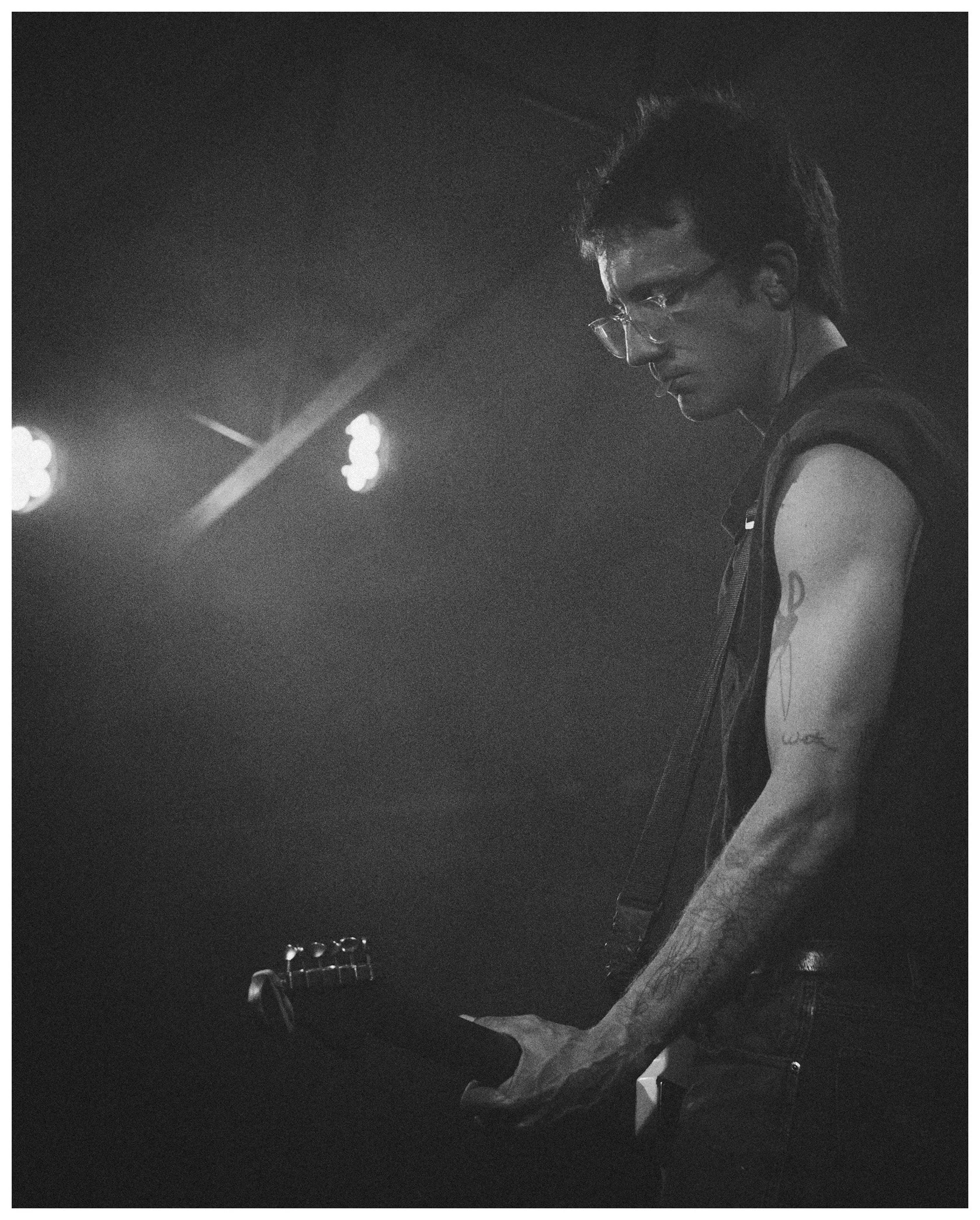 A person with dark hair and glasses playing an electric guitar on stage, wearing a sleeveless shirt, with stage lights in the background