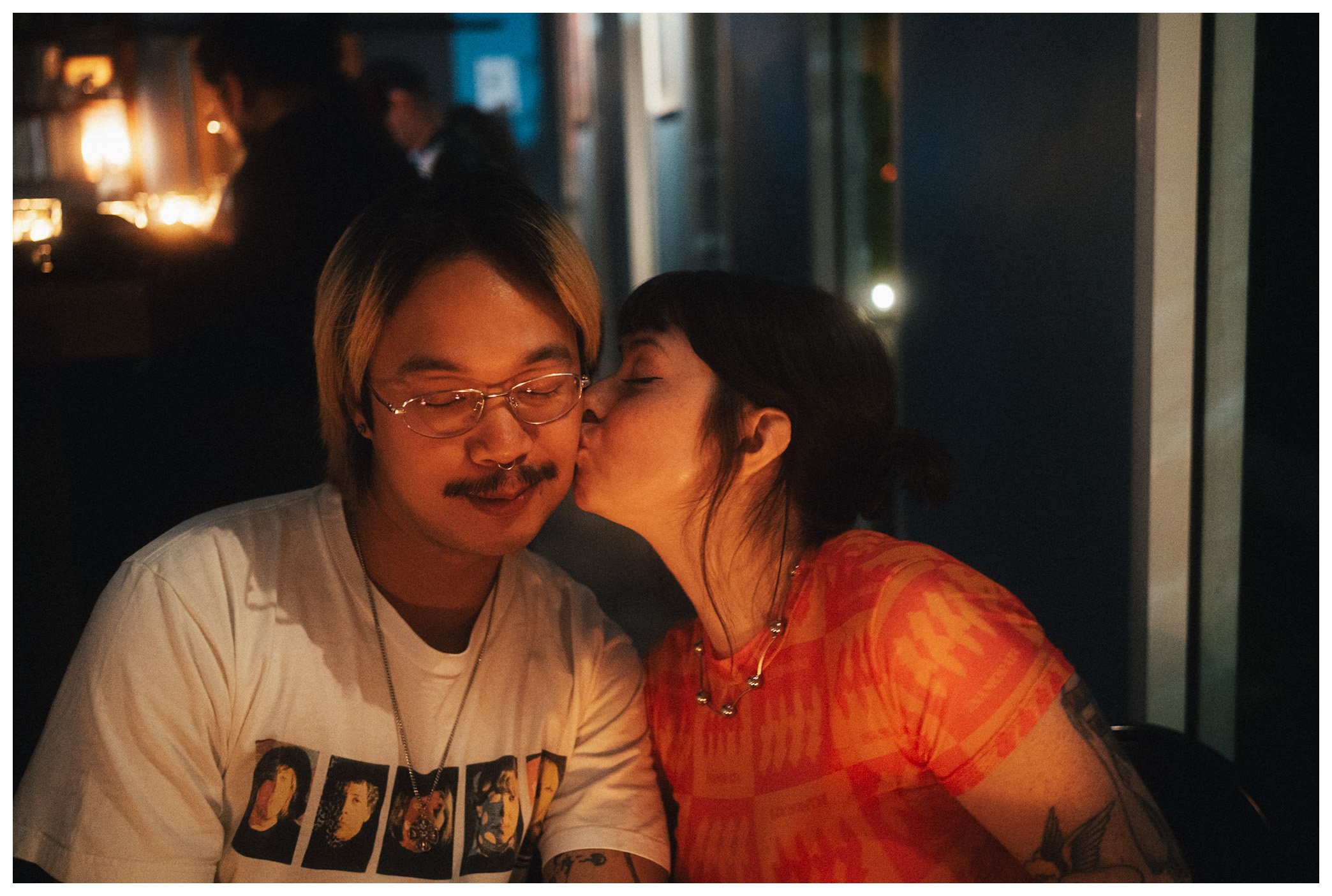 A man with glasses and a tattoo on his arm is smiling with his eyes closed as a woman on the right is giving him a kiss on the cheek. They are indoors in a dimly lit space, possibly a restaurant or cafe.