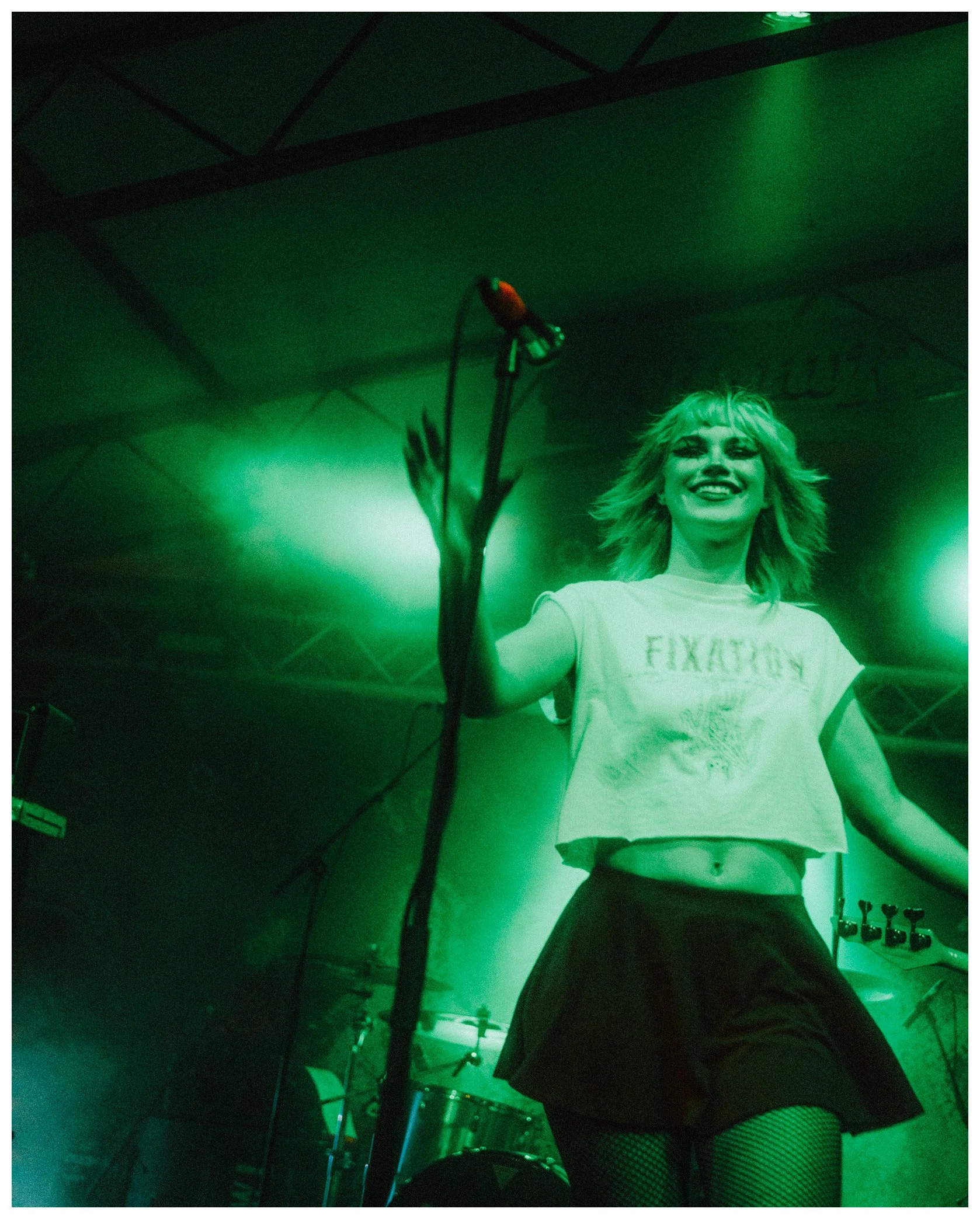 A woman with blonde hair and makeup smiling, wearing a white t-shirt and fishnet stockings, standing on a stage with green lighting, with a drum set partially visible at the bottom.