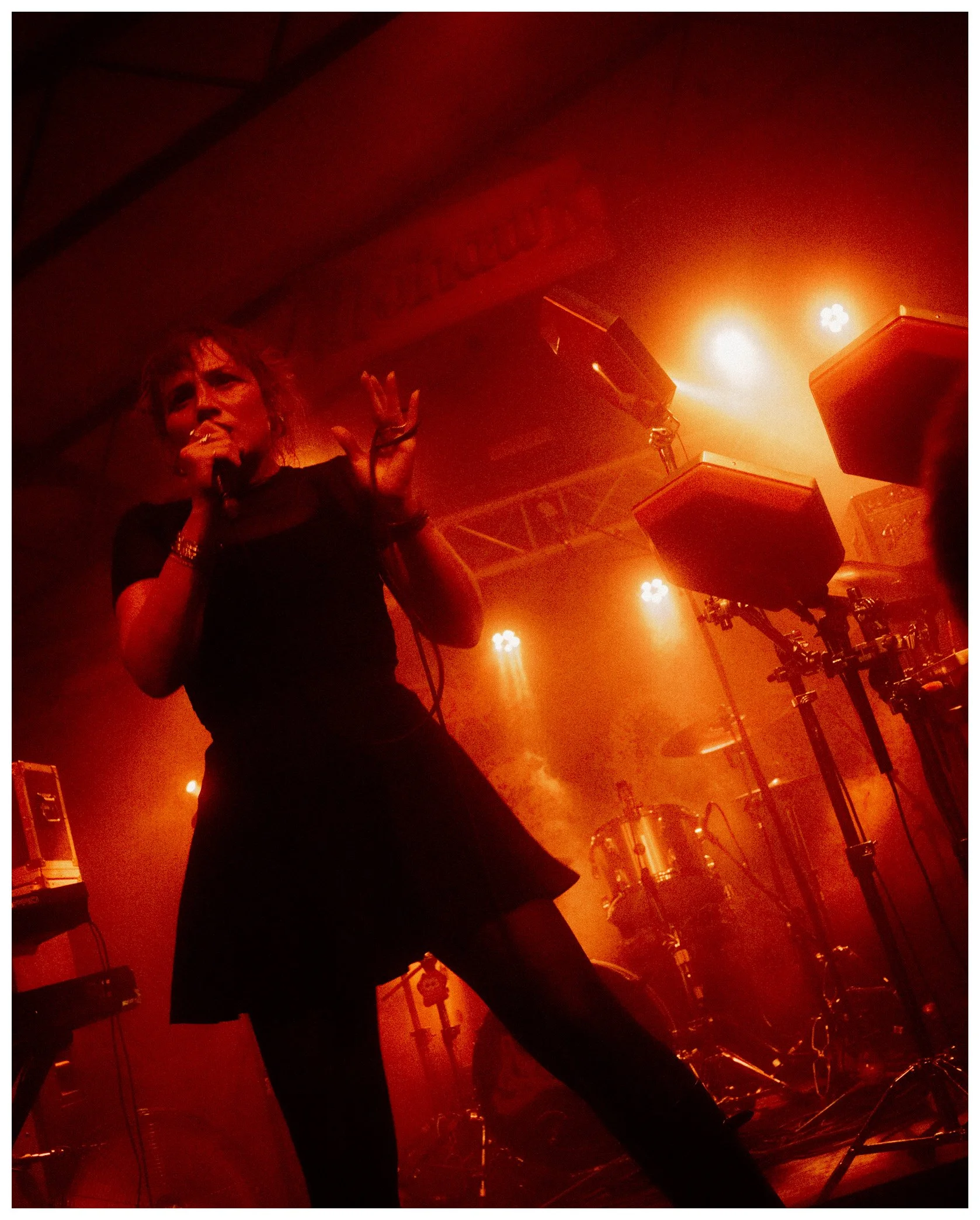 A woman with curly hair singing into a microphone on stage, illuminated by red stage lights, with musical equipment around her.