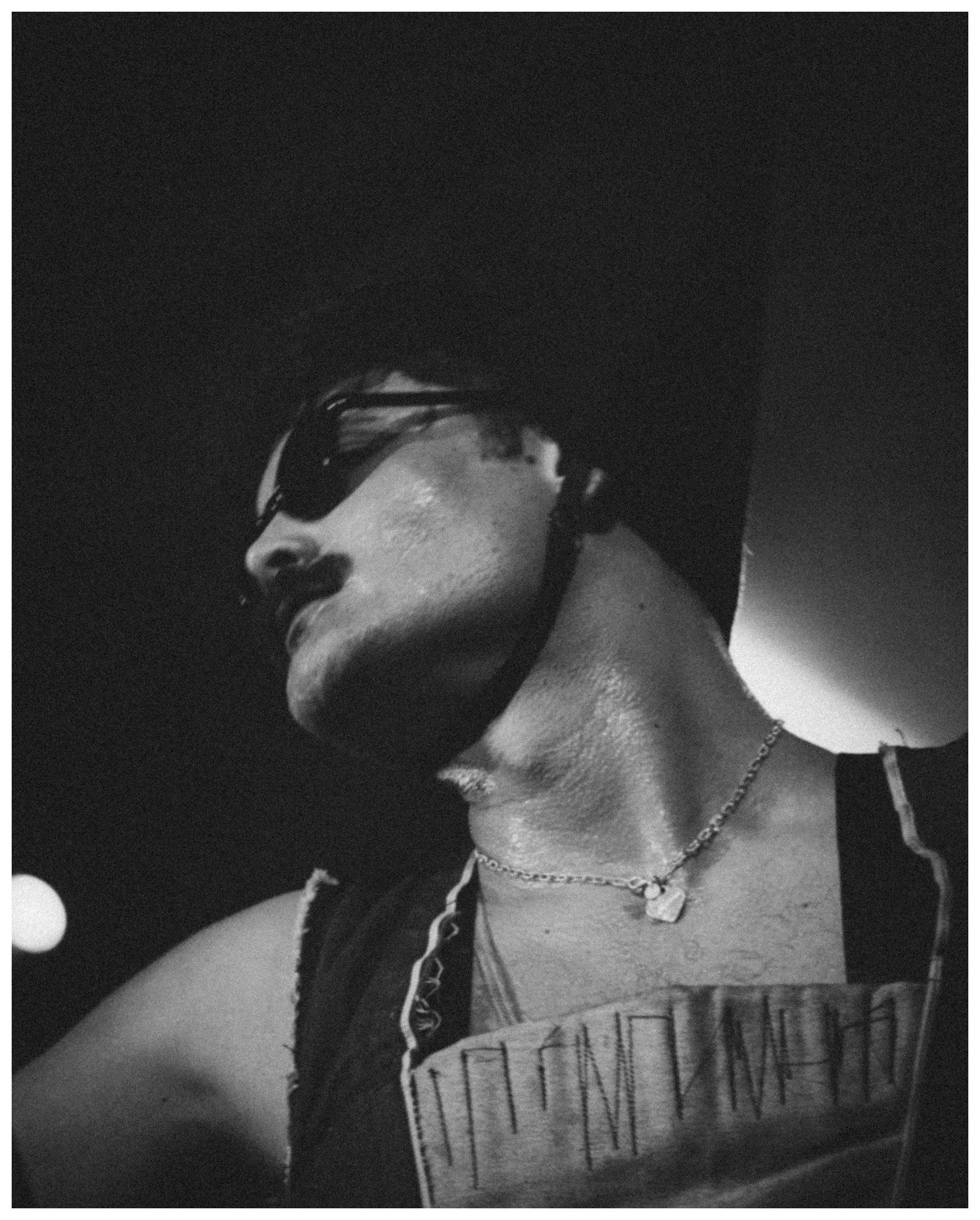 A black and white photo showing a woman wearing sunglasses and a necklace, with a notebook or journal with handwritten notes resting on her lap.