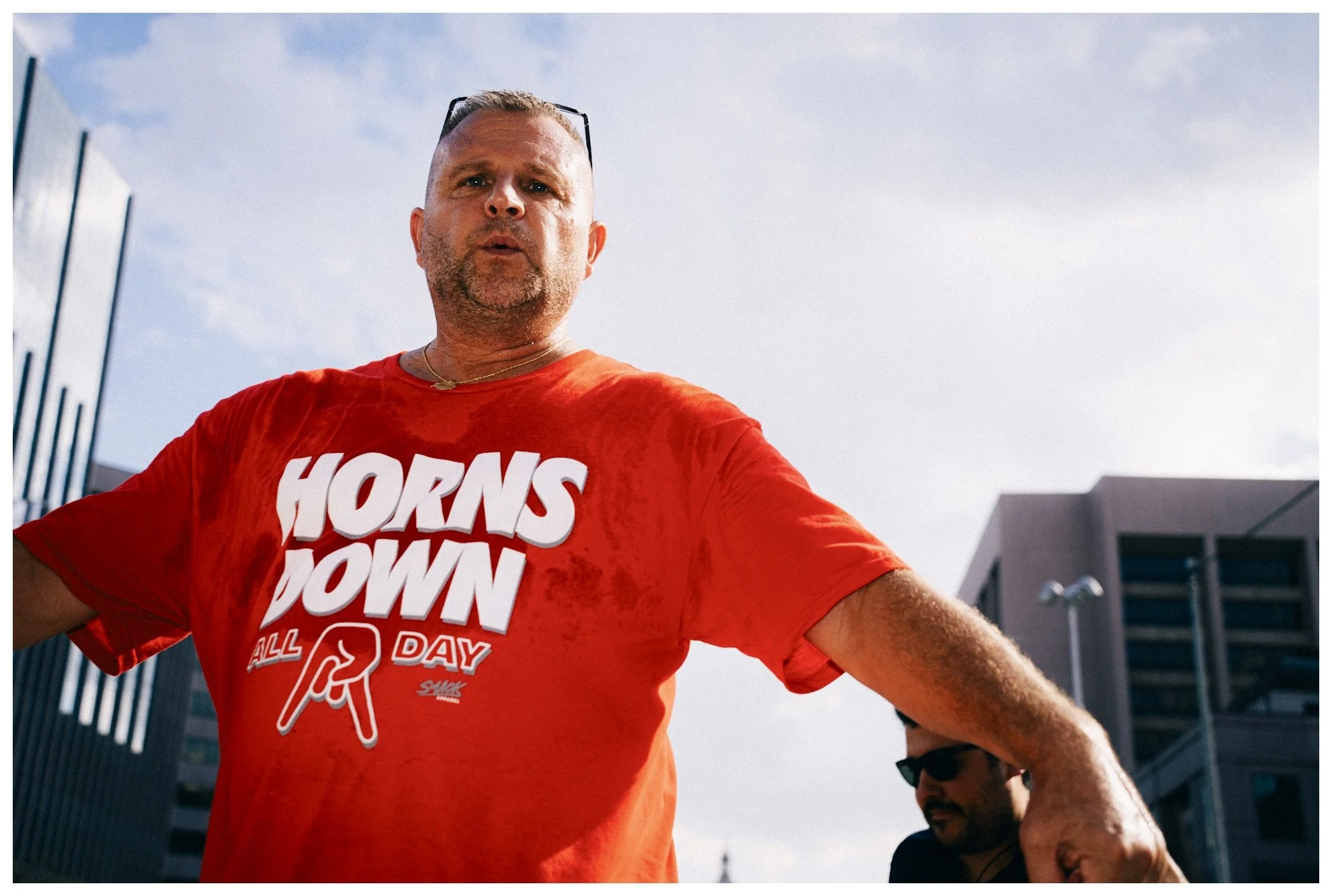A man wearing a red t-shirt with the text 'HORNS DOWN' on it, standing outdoors with a modern building and a partly cloudy sky in the background.