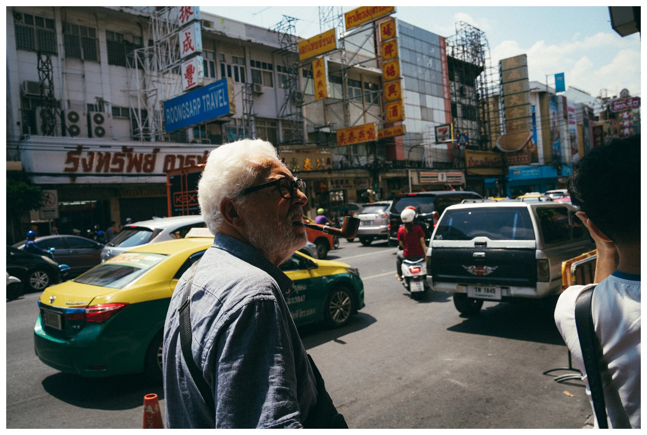 A man with white hair and a beard smoking a pipe stands on a busy city street with cars and motorcycles; neon signs and buildings in the background.