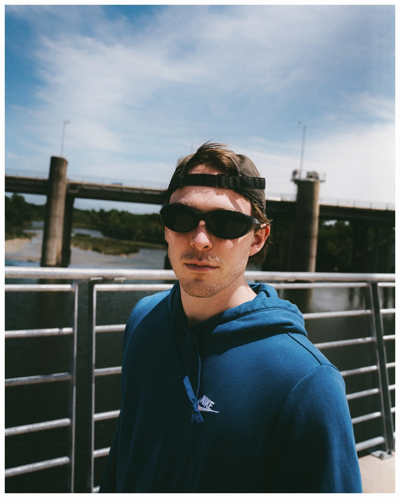 A person wearing a blue Nike hoodie, sunglasses, and a backward baseball cap, standing outdoors near a waterway with a bridge and green trees in the background.