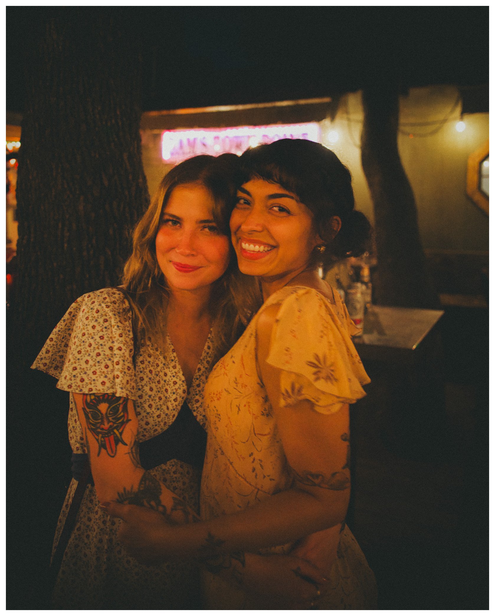 Two women smiling and taking a selfie in a dimly lit environment, possibly a bar or club, with a neon sign in the background.