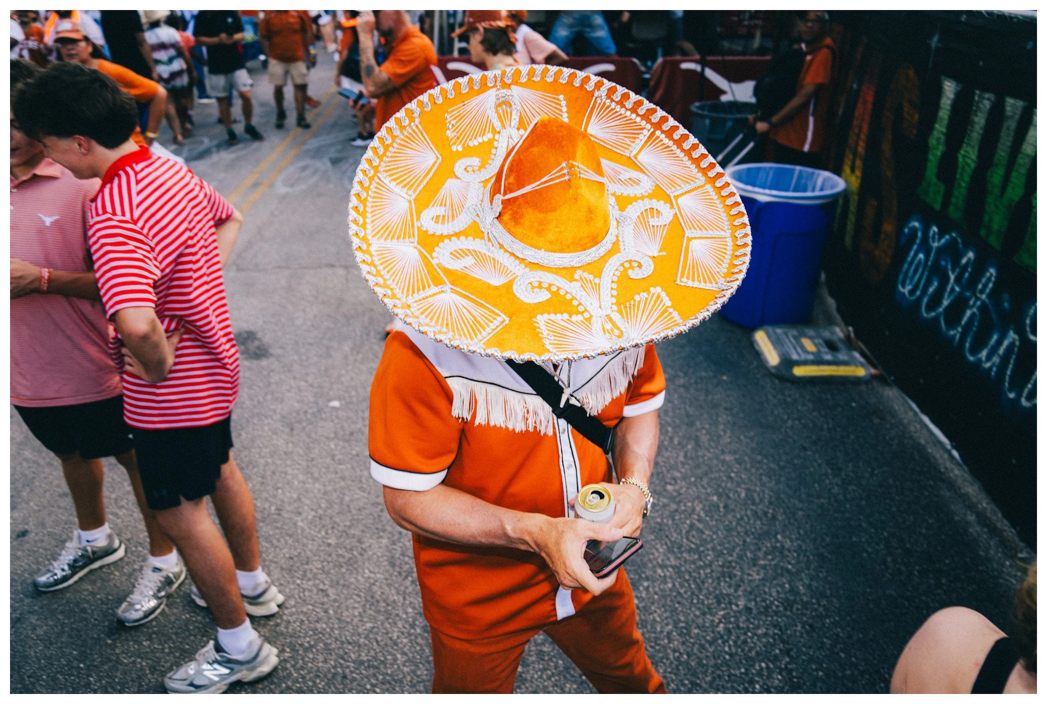 Person wearing a bright yellow sombrero with white decorative design at a street event. The person is dressed in an orange outfit and holding a beverage can and phone. There are people in casual attire around, some playing and socializing.