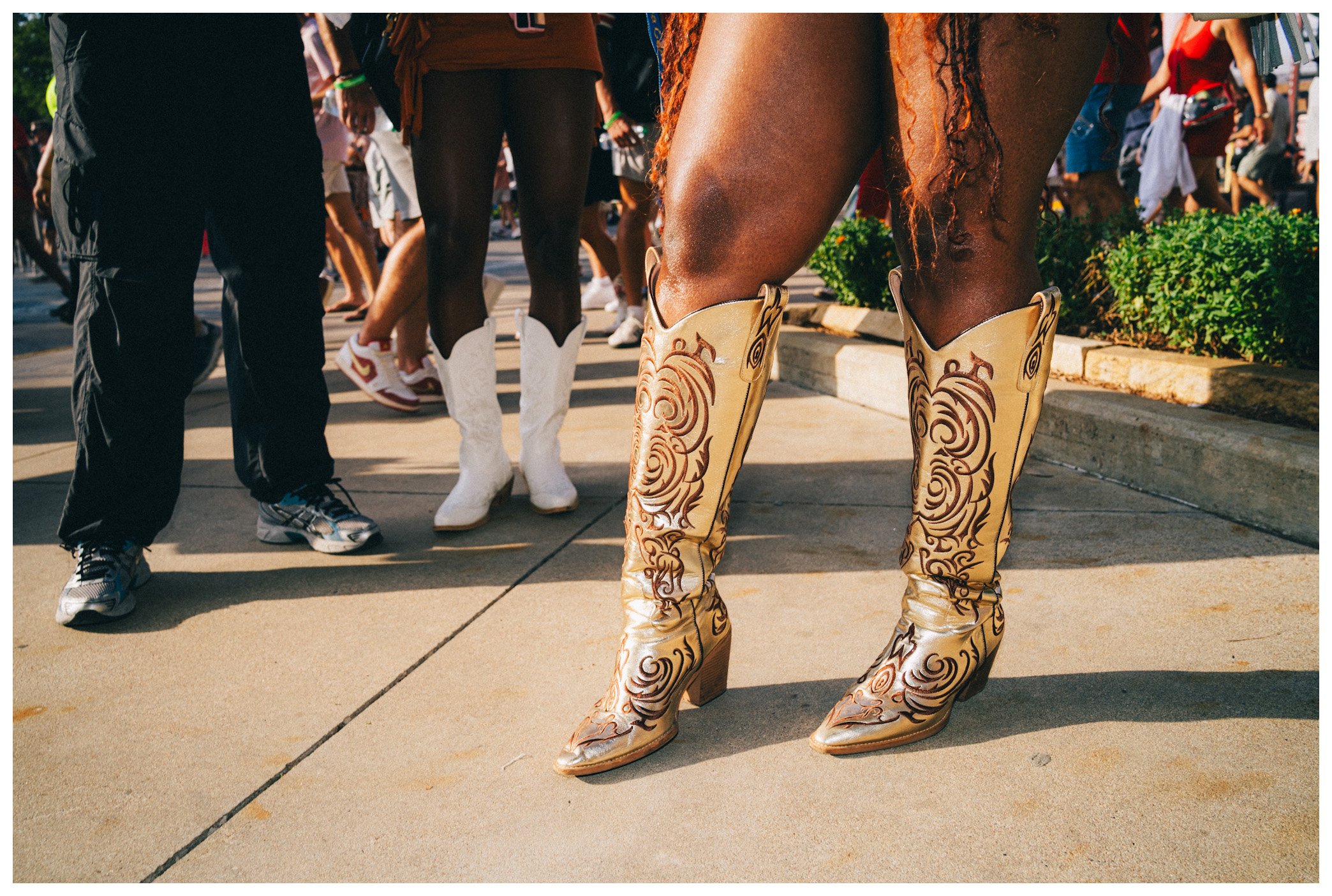 Close-up of a woman wearing tan cowboy boots with elaborate dark brown swirling designs, standing among a crowd of people outdoors.