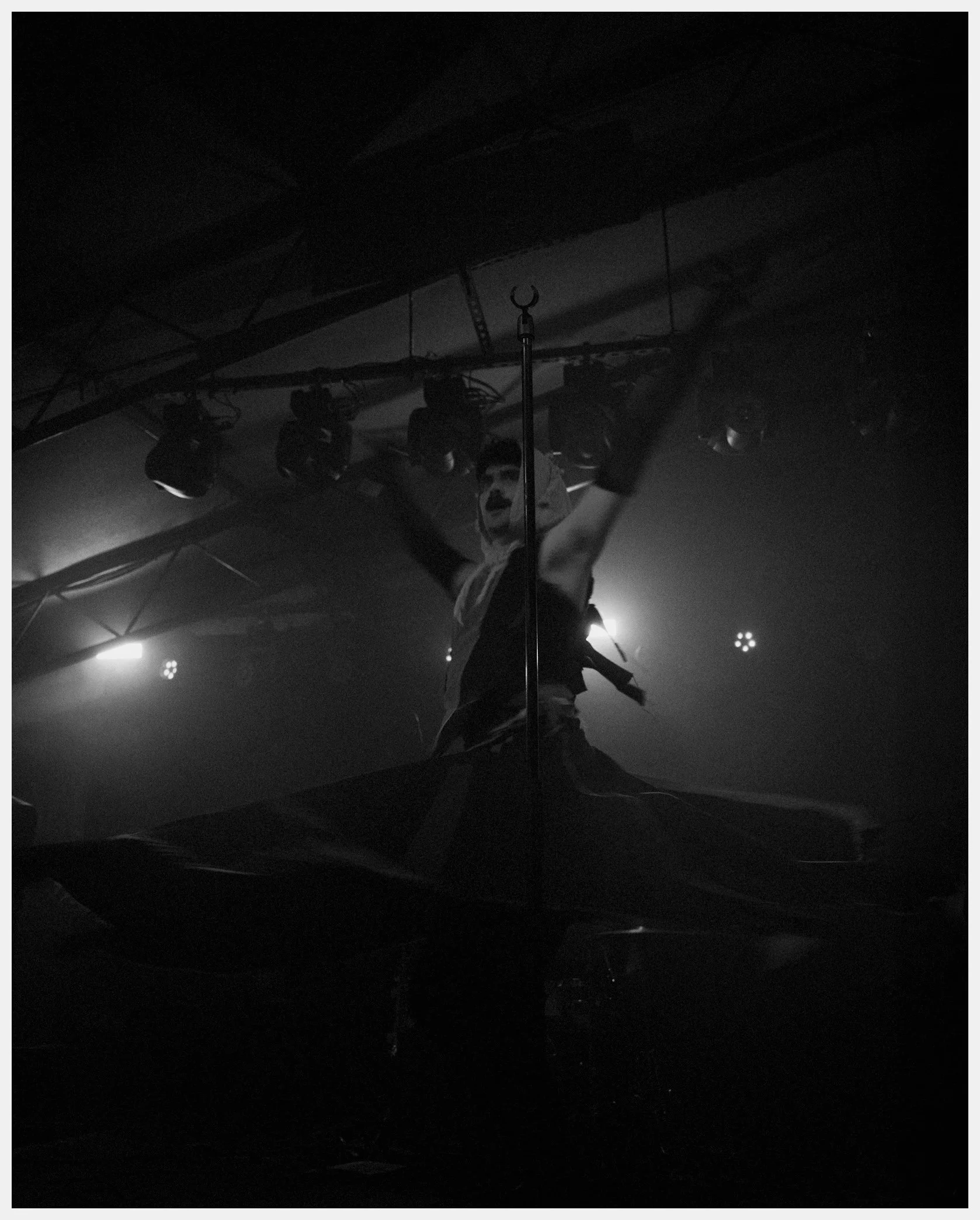 Dark indoor scene with stage lighting, a person standing under stage lights, holding objects, with visible stage and metal truss overhead, in a dimly lit environment.