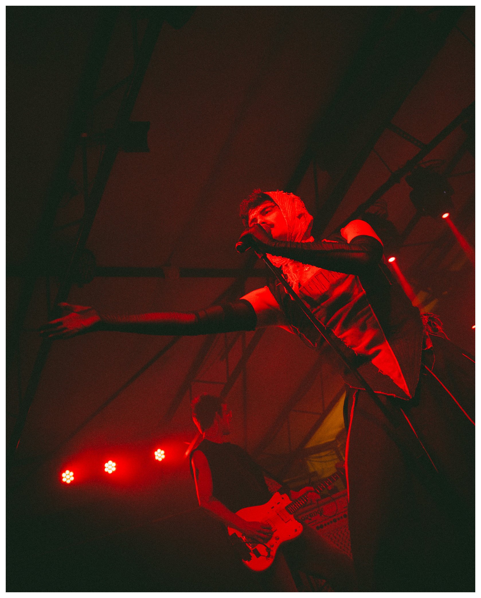 A performer on stage illuminated by red lights, holding a microphone and wearing a red hooded jacket with patterns.