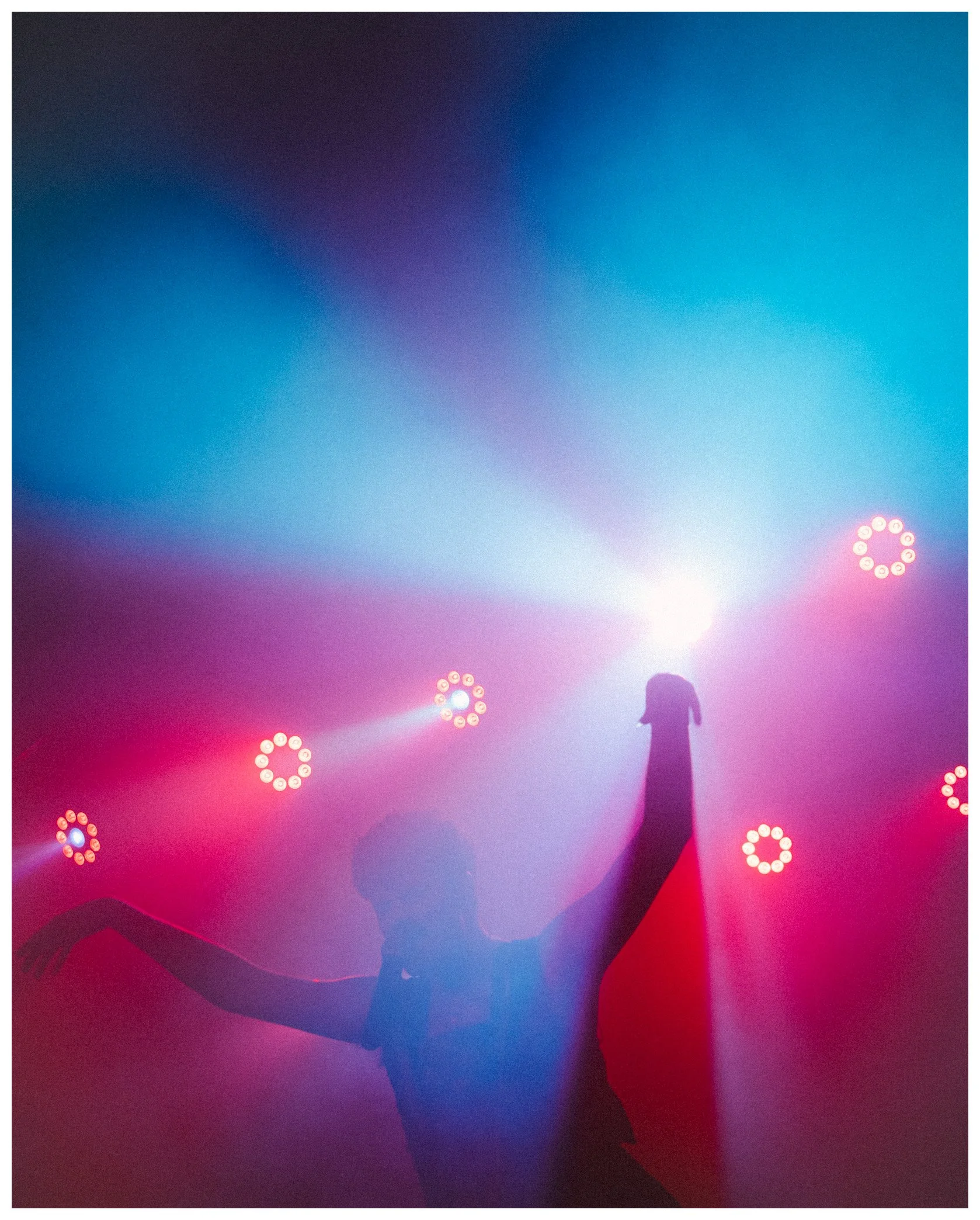 Silhouette of a person singing or speaking into a microphone on stage, with pink and purple lighting and audience or people in the foreground.