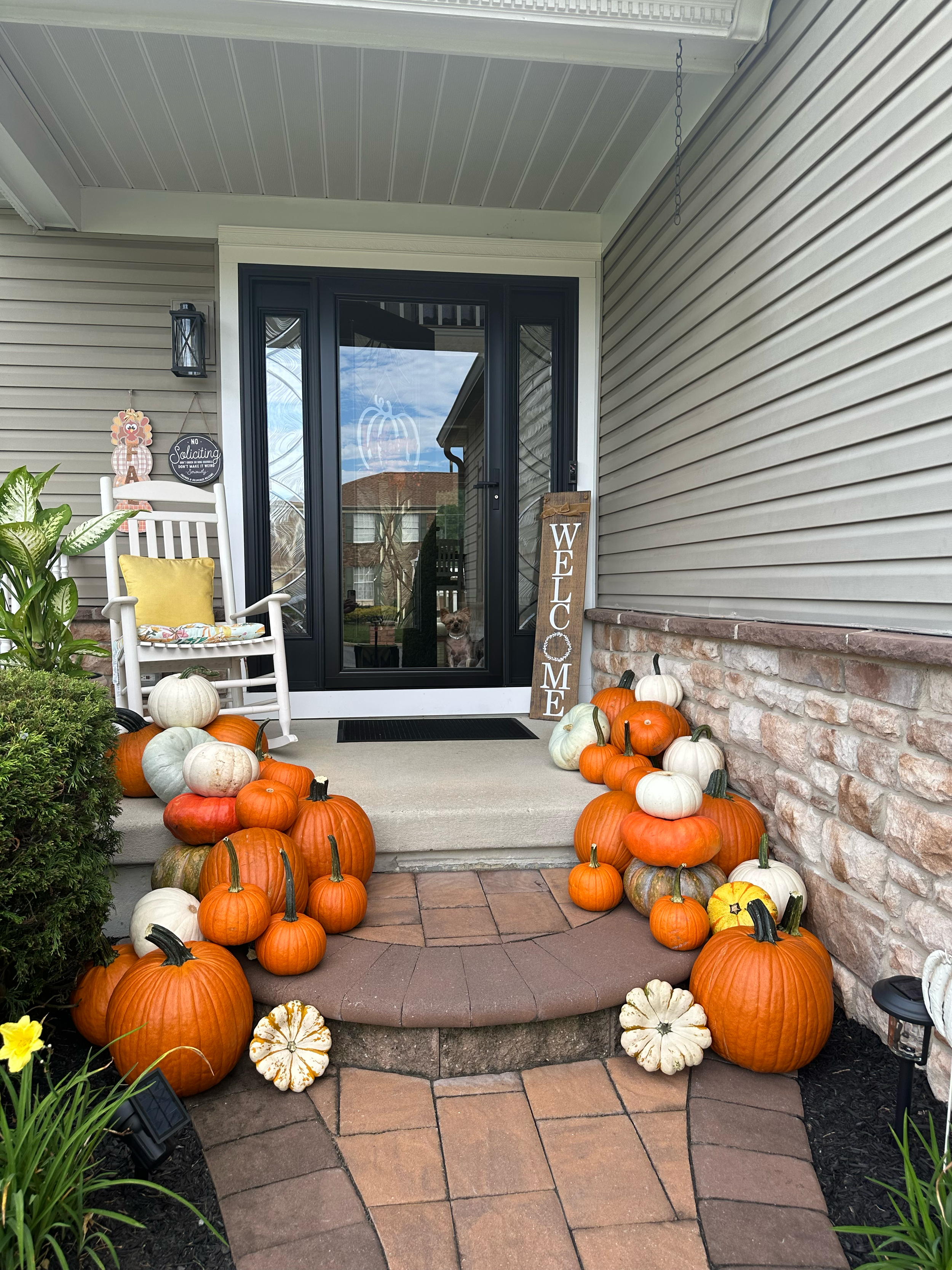 Front porch decorated for fall with numerous pumpkins of various sizes and colors, a white rocking chair with a yellow cushion, a welcome sign, and fall plants.