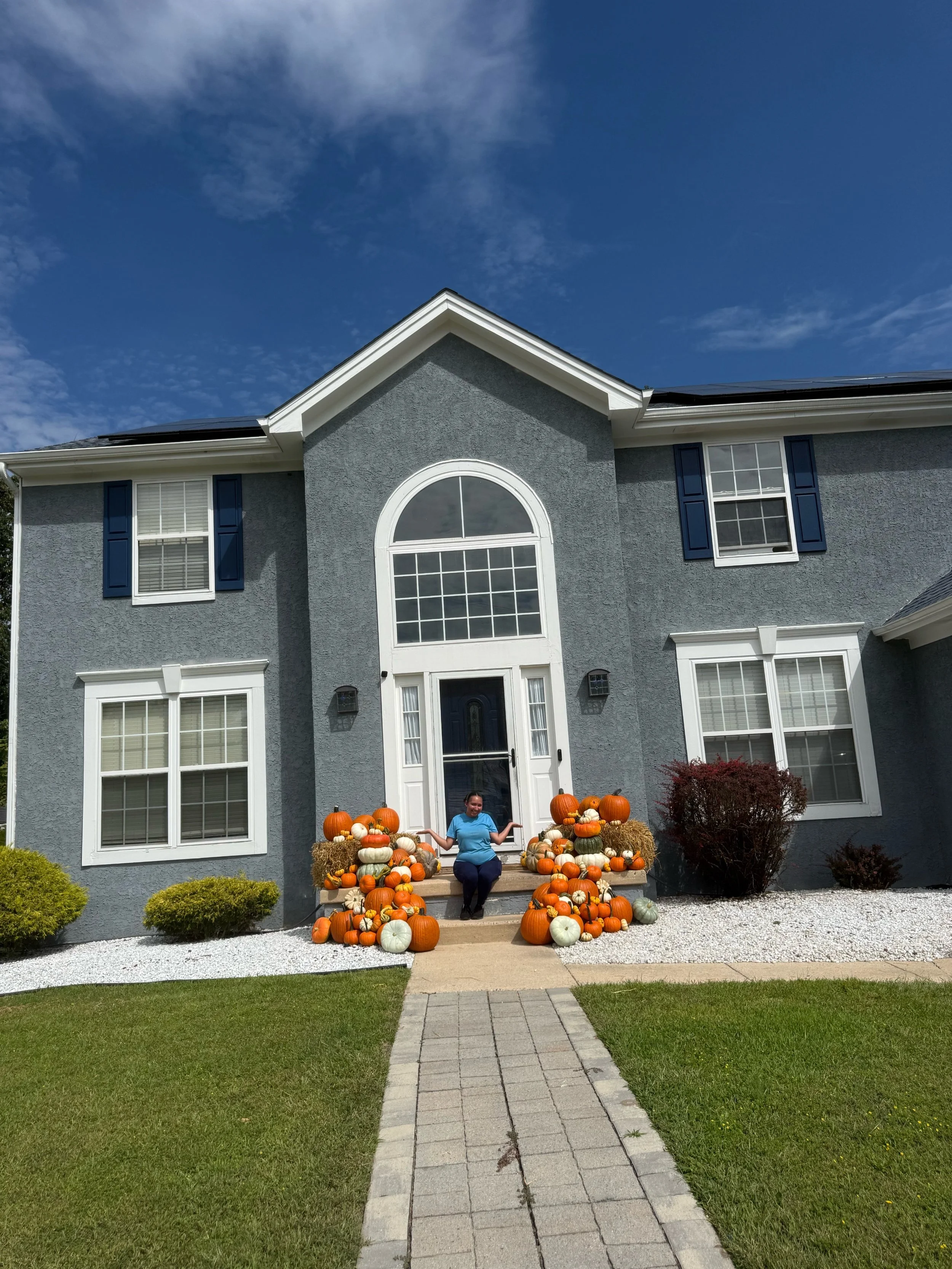 Woman sitting beside a fall pumpkin display arranged on the steps of a light blue home.