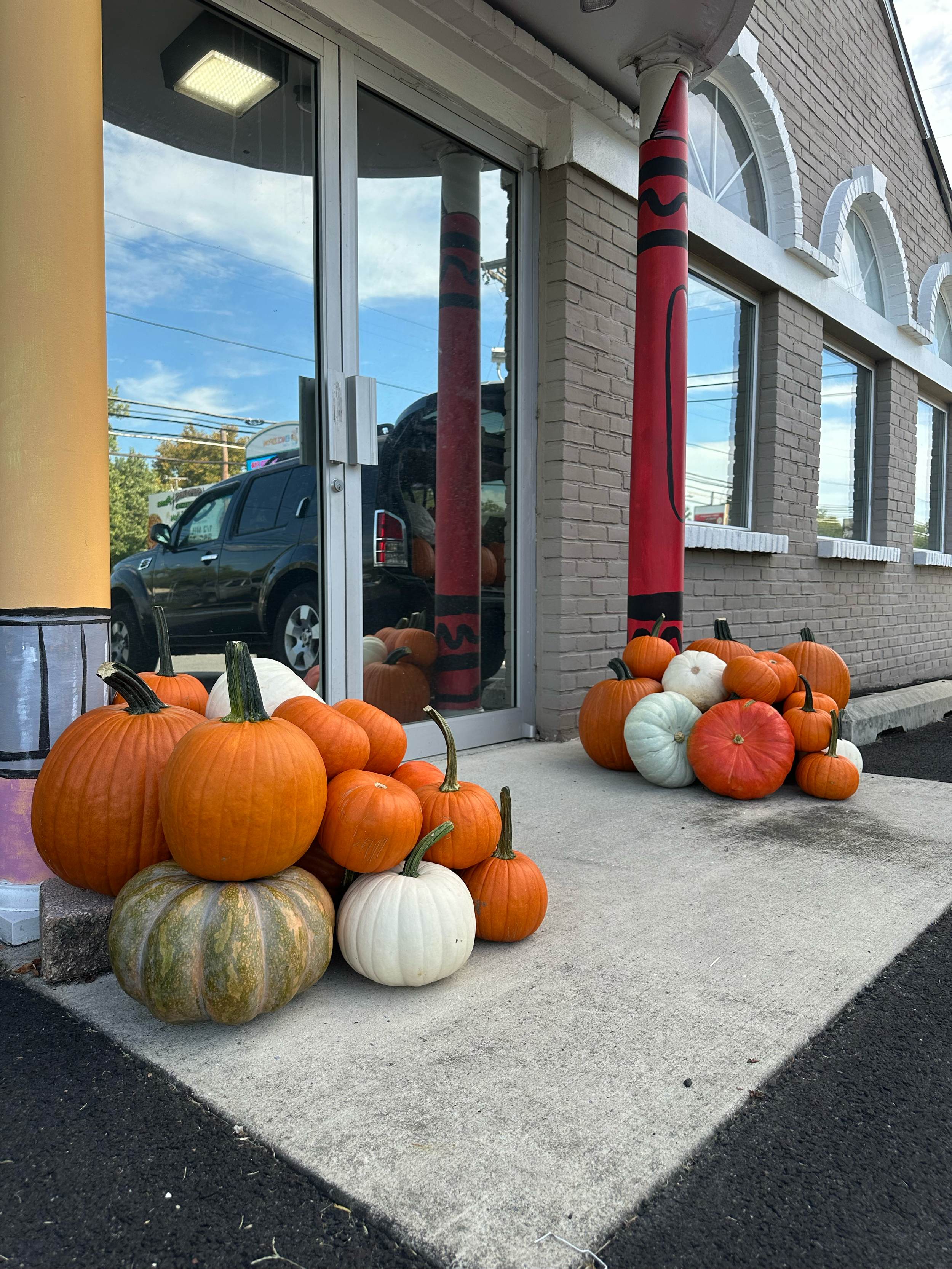 A group of pumpkins in various orange, white, green, and cream colors are arranged outside a building on a concrete sidewalk. The building has large windows with arched white trim and brick walls. Two red and black striped poles are positioned between the pumpkins and the building. 