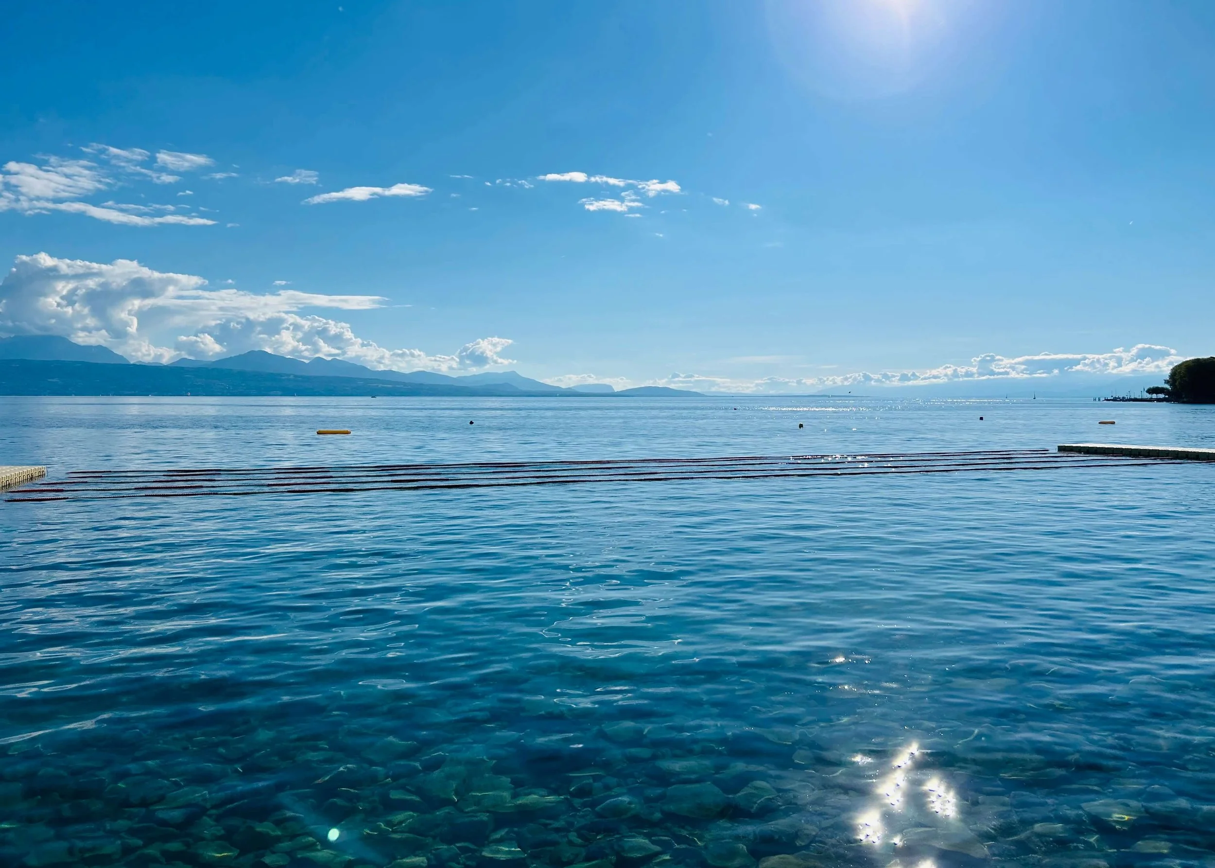 Photo du Léman calme avec un ciel bleu clair et quelques nuages, ainsi que des montagnes visibles à l'horizon.