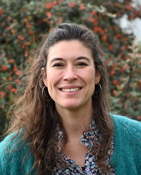 Emilie Stein, Psychologue FSP, formée aux TCC, MBSR, ACT, auto-compassion, souriante avec des cheveux bouclés brun, portant une veste bleue et un chemisier à motifs, en extérieur avec un arbuste aux feuilles rouges en arrière-plan.