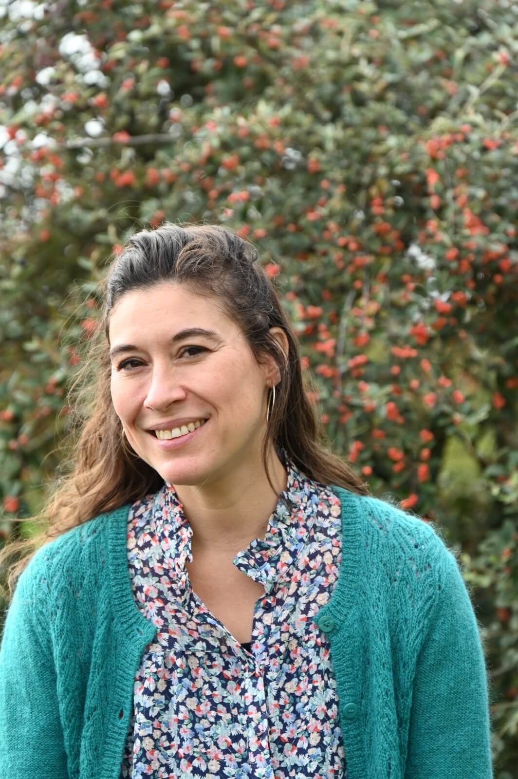 Jeune femme souriante avec des cheveux longs, portant un pull turquoise et une chemise à fleurs, posant devant un buisson aux petites baies rouges en arrière-plan.