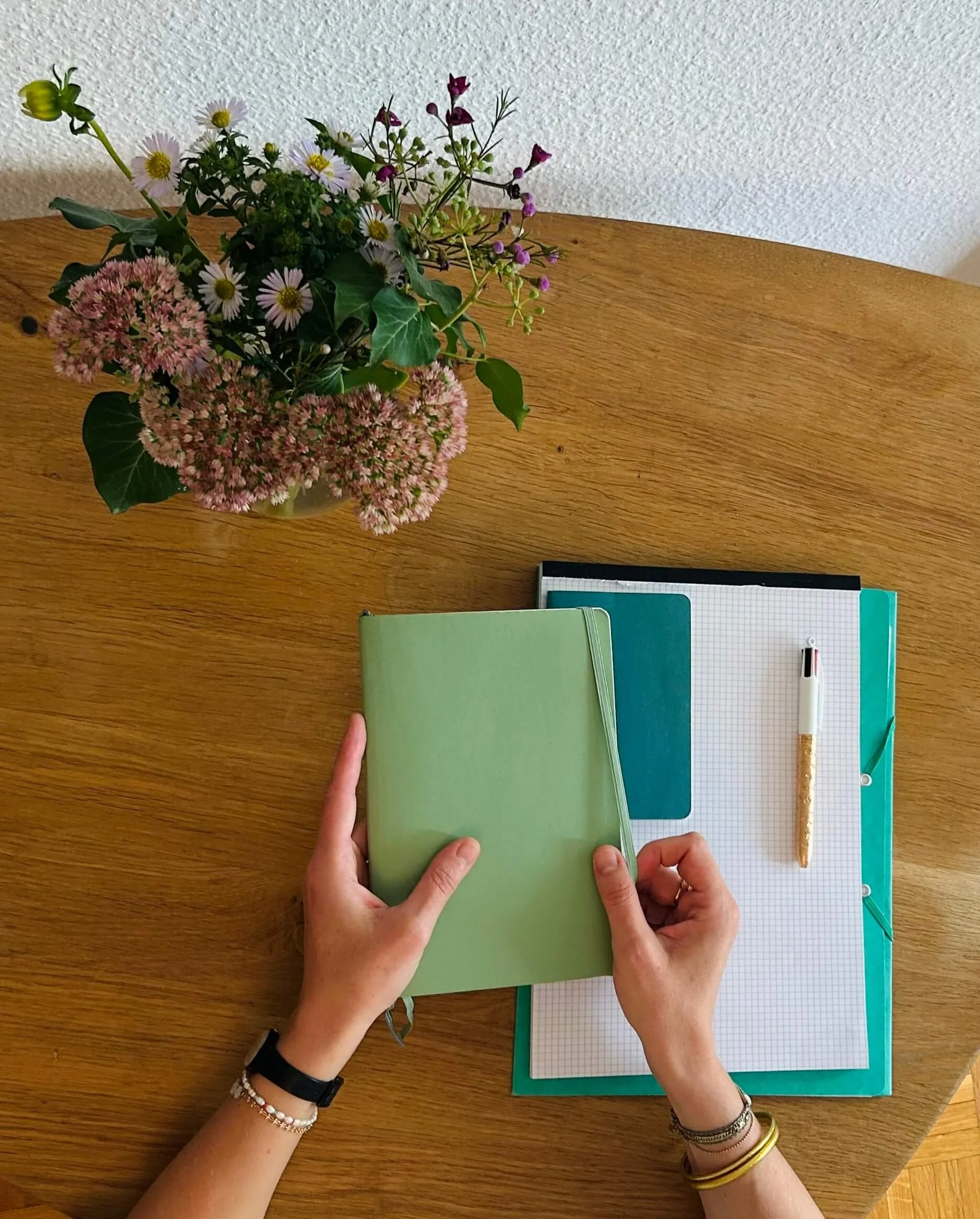 Emilie Stein, psychologue FSP à Lausanne, tient un cahier vert avec deux mains, un stylo à côté, sur une table en bois, au-dessus d'un bouquet de fleurs dans un vase et de carnets.