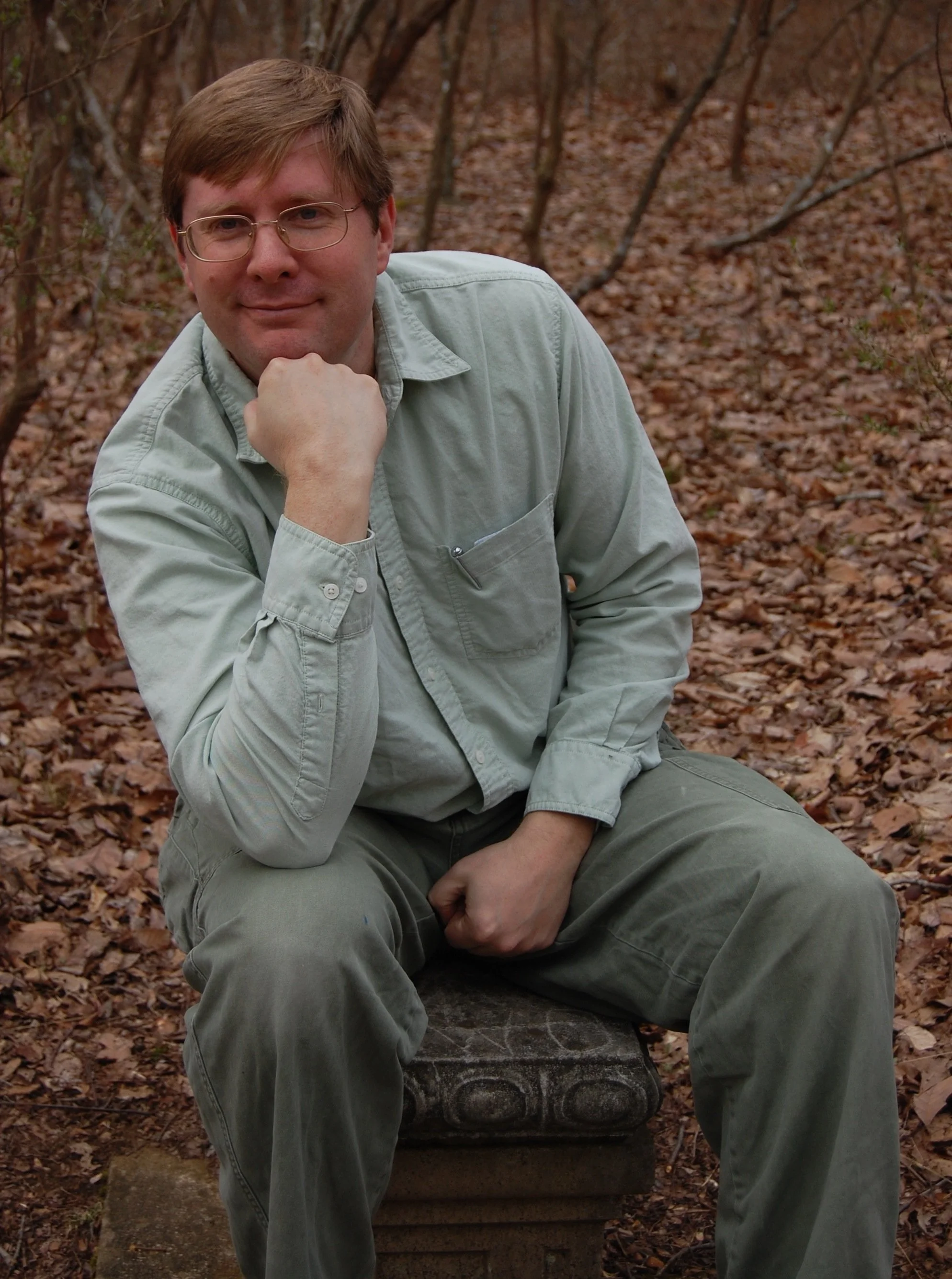 A photo of David Young in a thinker pose sitting on a plinth in a wooded area. He was in his fourties in the picture.