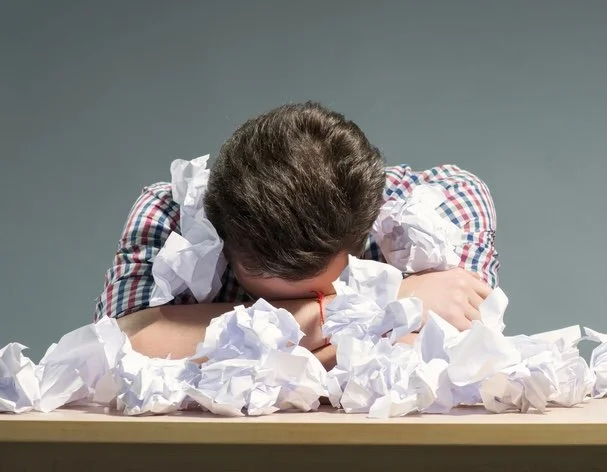 A picture of a man with his head down surrounded by crumpled pieces of paper