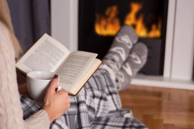 Cozy image of a person reading a book and holding a mug in front of a fire.