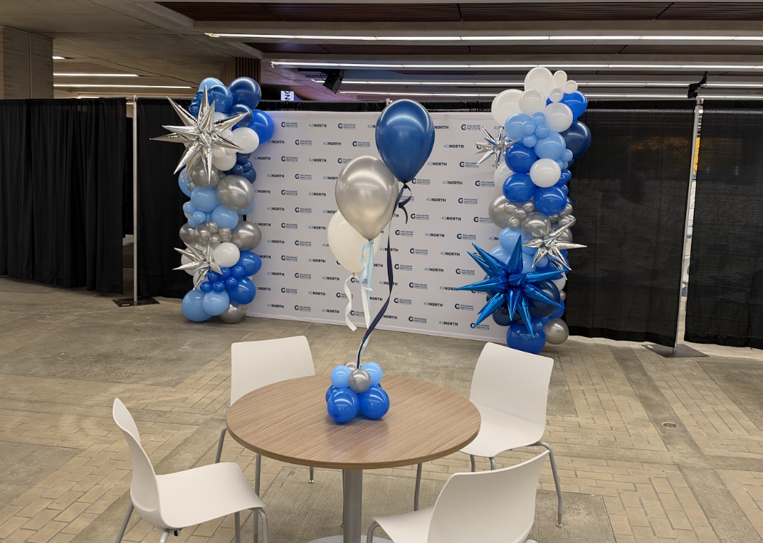 Balloon arrangement with blue, white, silver, and star-shaped balloons on a table in front of a backdrop with logos, set up for an event or celebration.