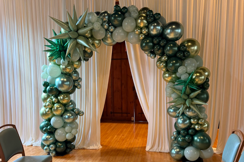 Balloon arch with gold, silver, and white balloons, decorated with large green metallic stars, set in front of cream curtains and wooden doors.
