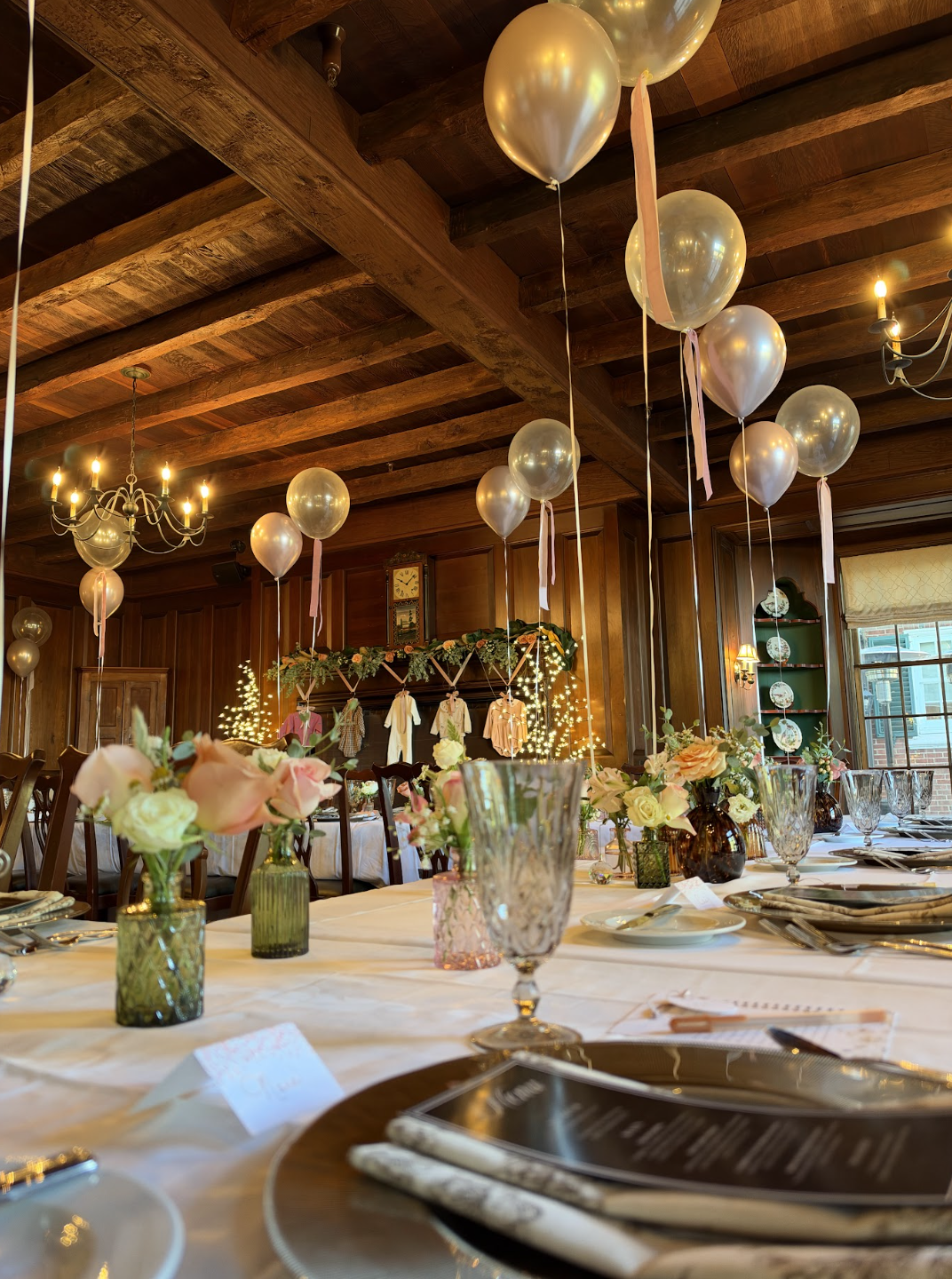 Decorated banquet hall with pink and white balloons hanging from the ceiling, floral centerpieces on tables, and festive string lights in a cozy wood-paneled room.