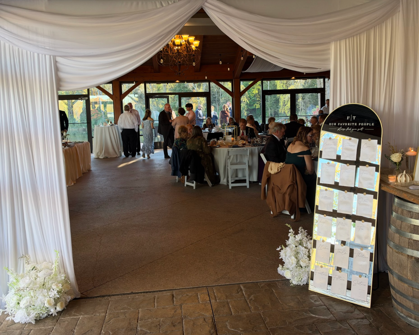 Indoor wedding reception with guests seated at round tables, some standing and chatting near large windows overlooking greenery, decorated with white drapes, flowers, and a display board with seating arrangements.